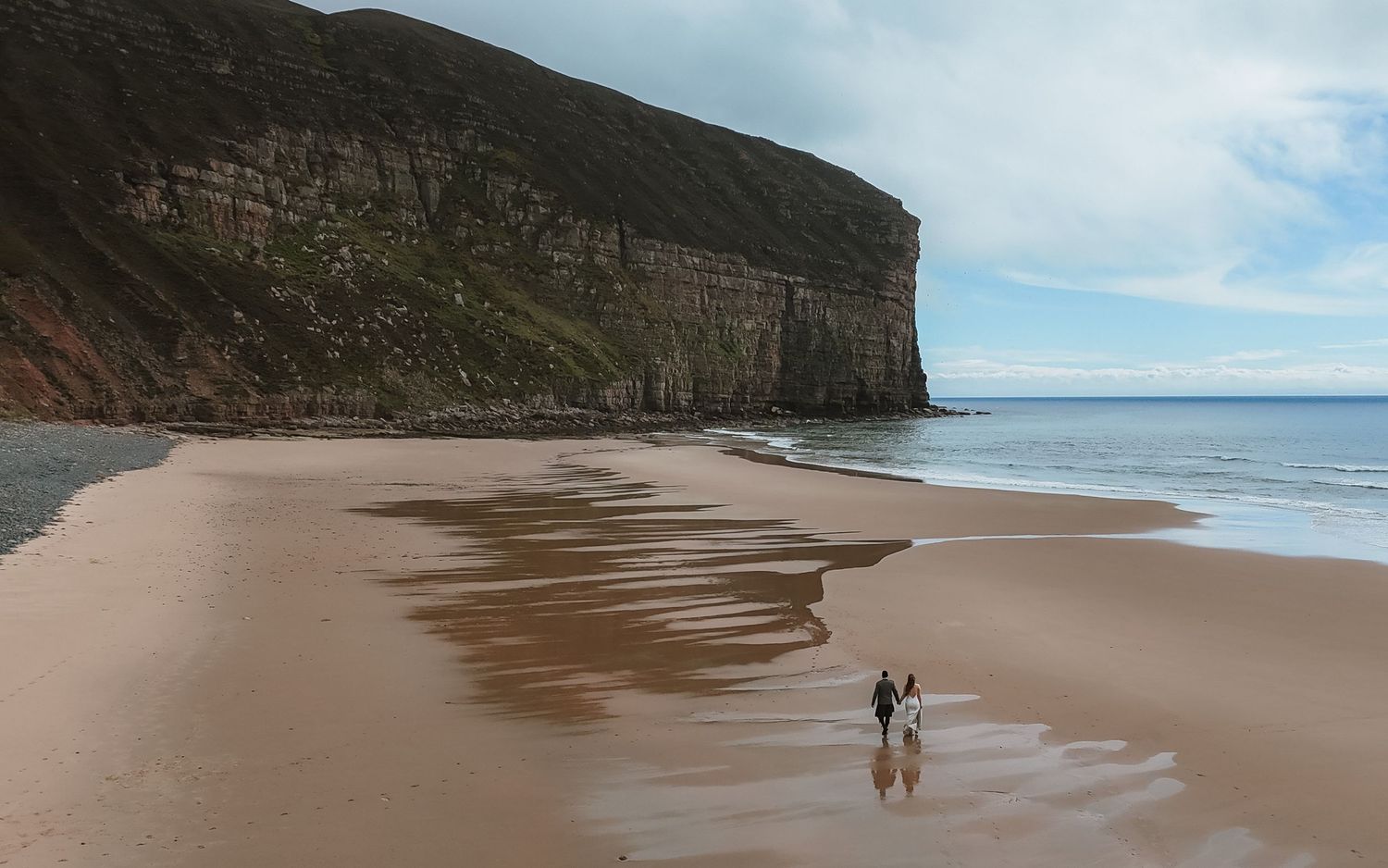 Two people walking along a vast sandy beach with towering cliffs and ocean waves under blue skies in Scotland.