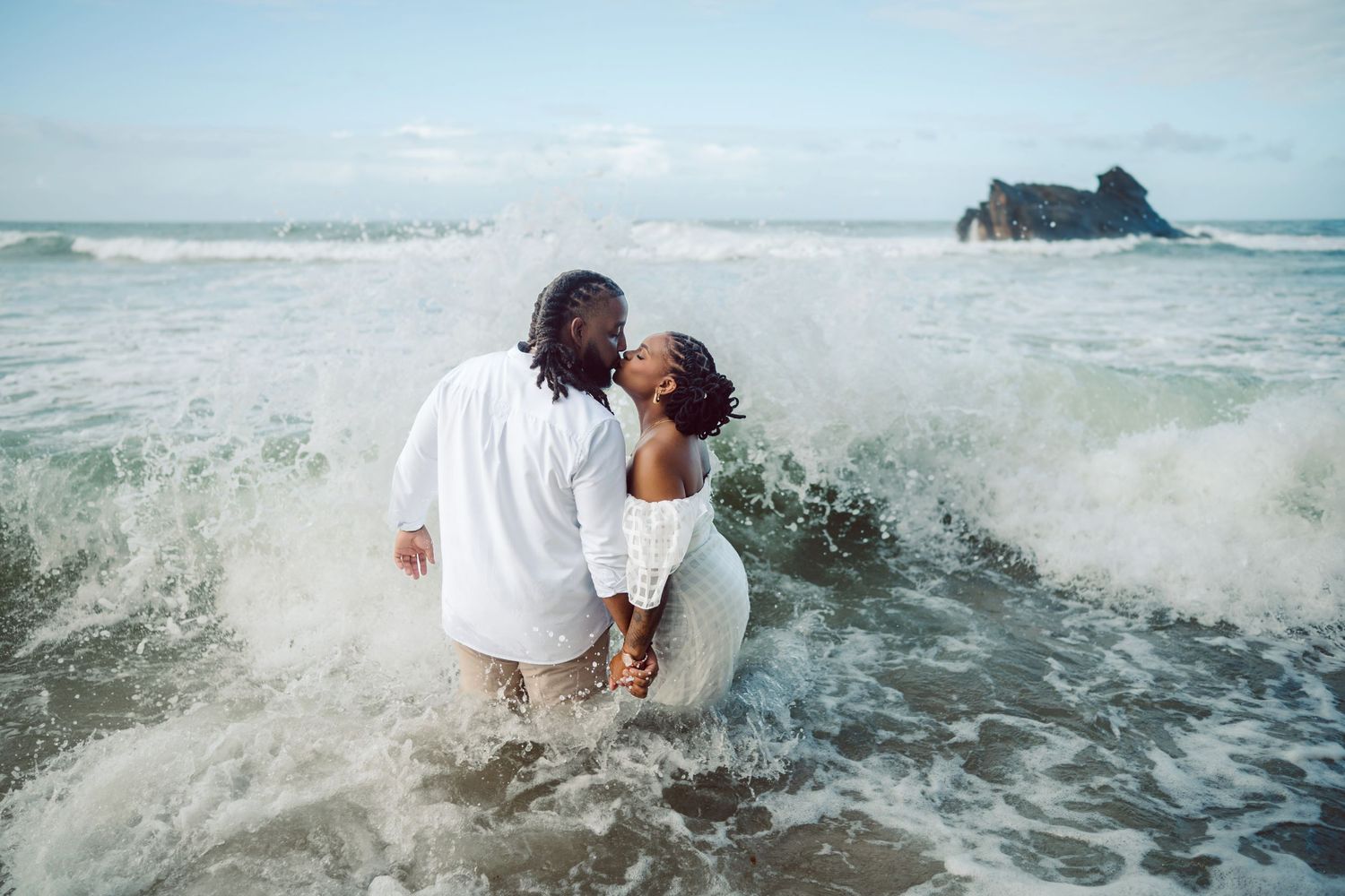 A couple shares an intimate moment at the beach as waves crash around them, with a rocky outcrop visible in the distance.