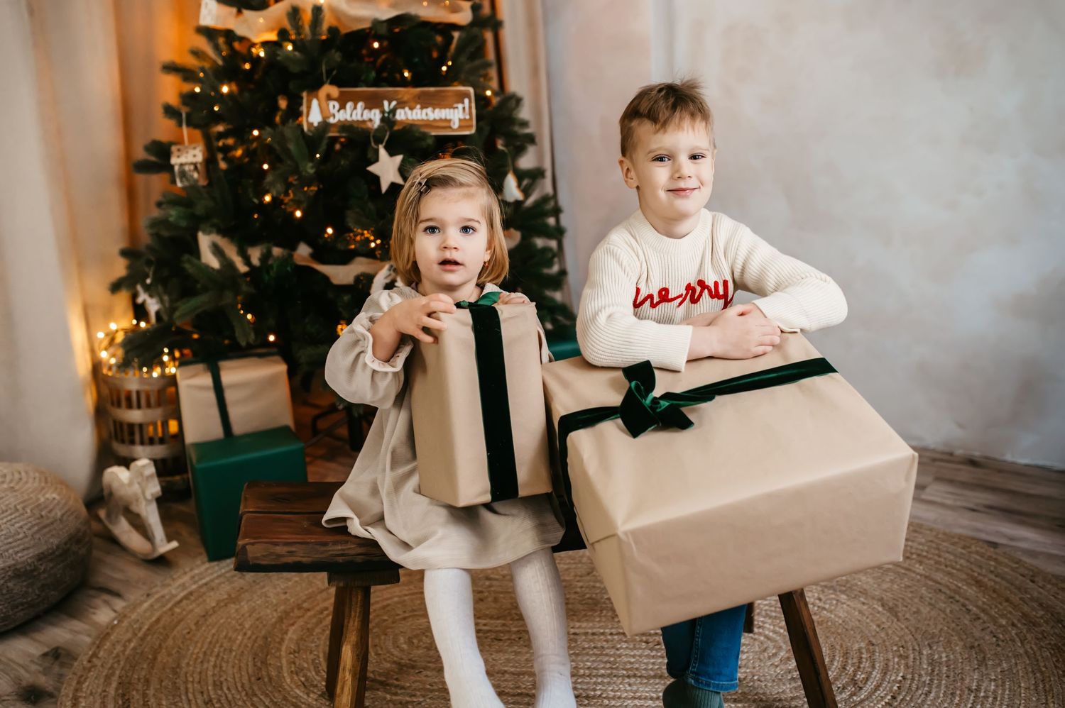 Two children sit with Christmas presents in front of a decorated tree with soft bokeh lights in the background.
