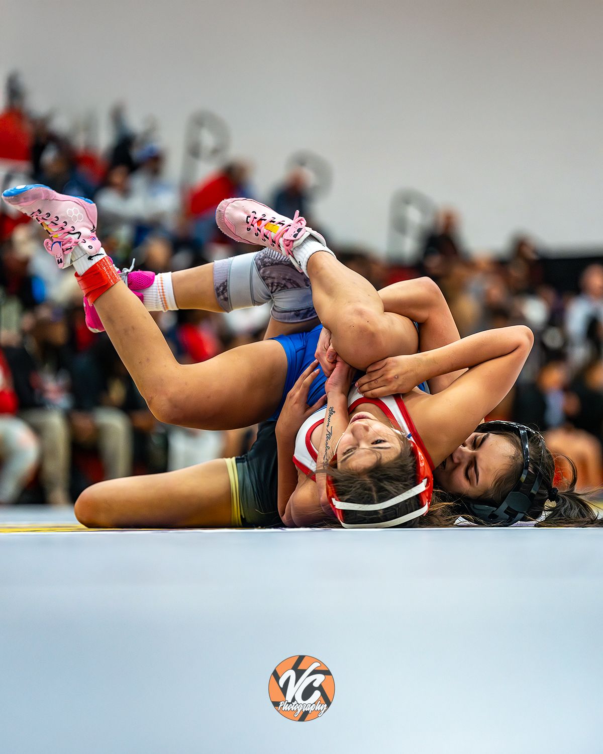 Two female wrestlers compete on a mat, one executing a takedown move during an indoor wrestling tournament.