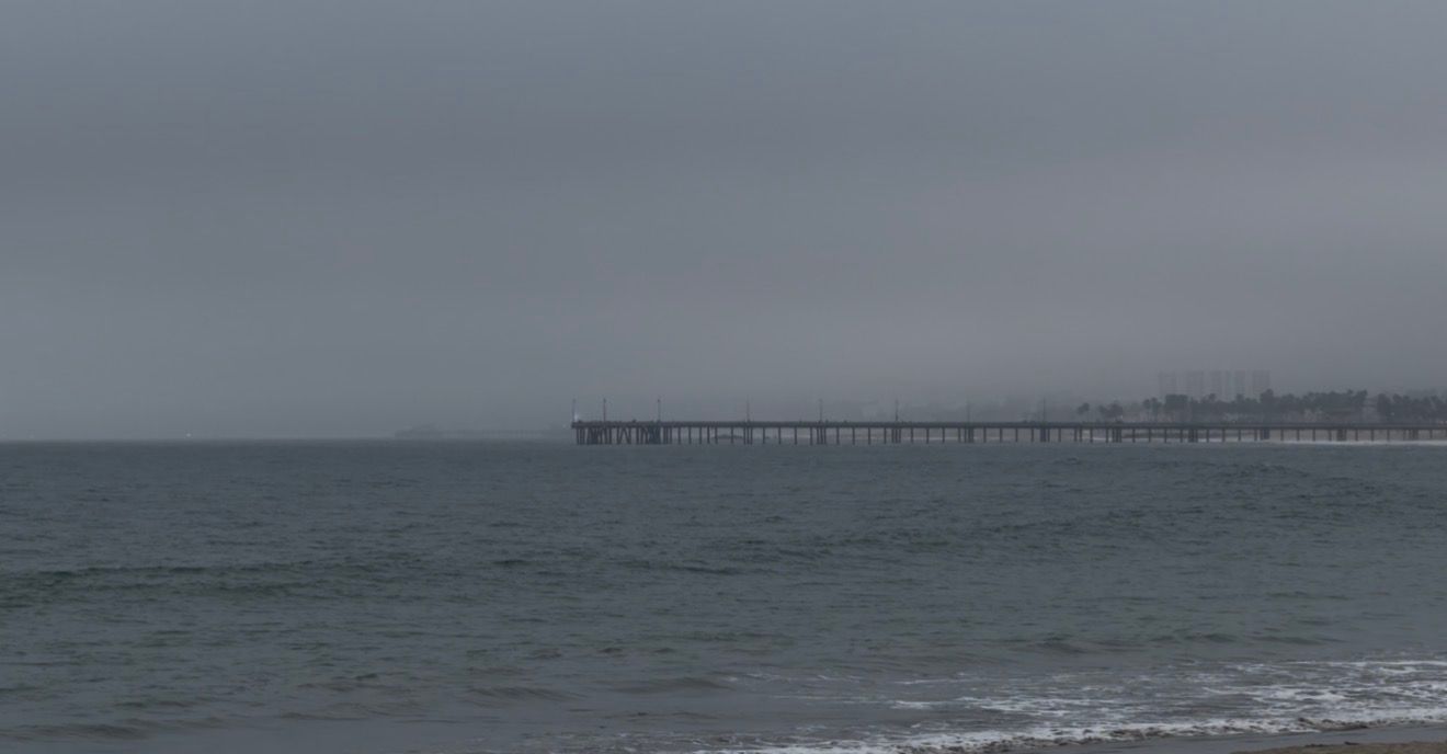 A long pier extends over calm gray ocean waters under an overcast sky on a cloudy day at the beach.