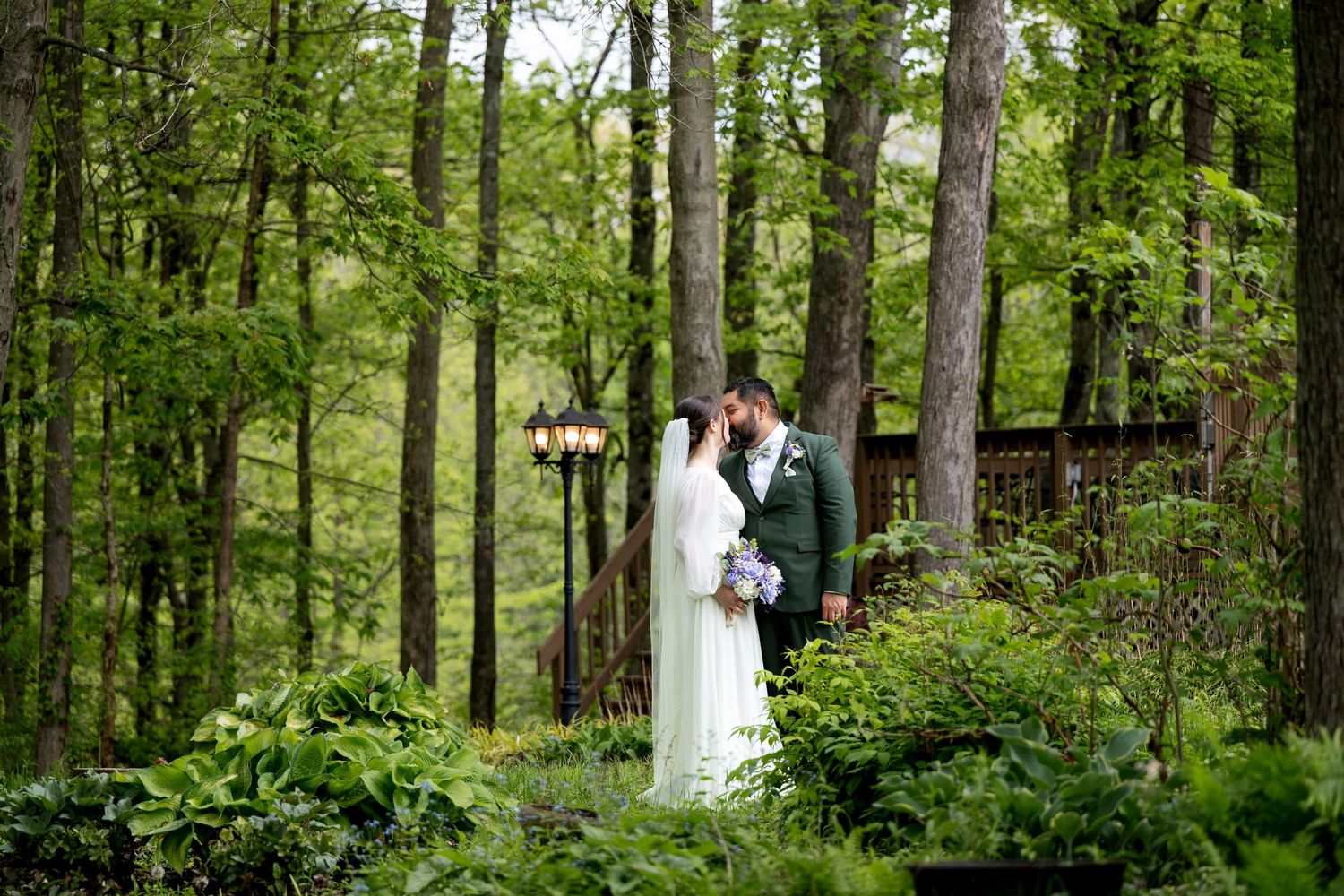 Bride and groom share a romantic kiss surrounded by lush green forest trees at an outdoor woodland wedding venue.
