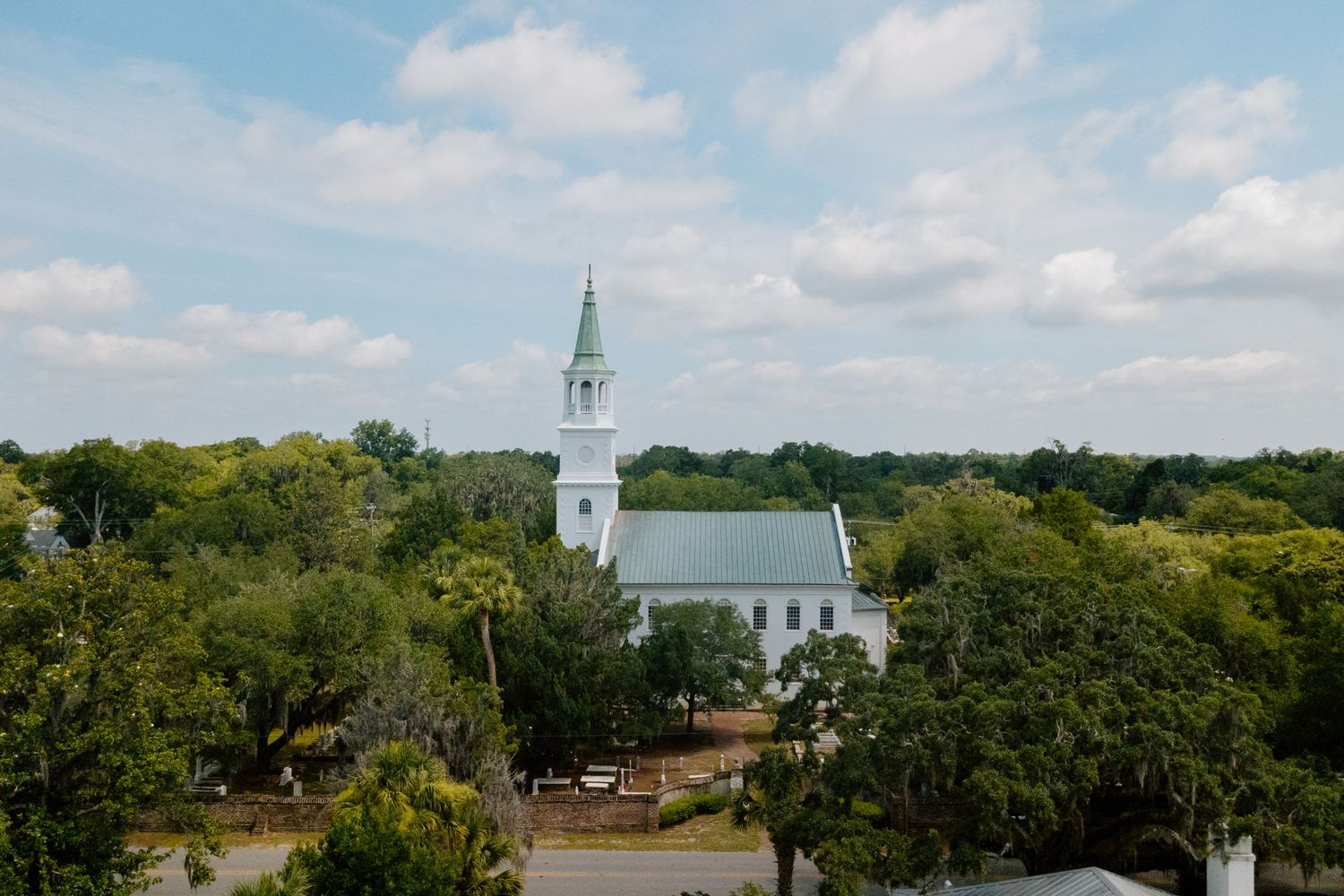 Aerial view of a white historic church with steeple surrounded by lush green trees under a partly cloudy sky.