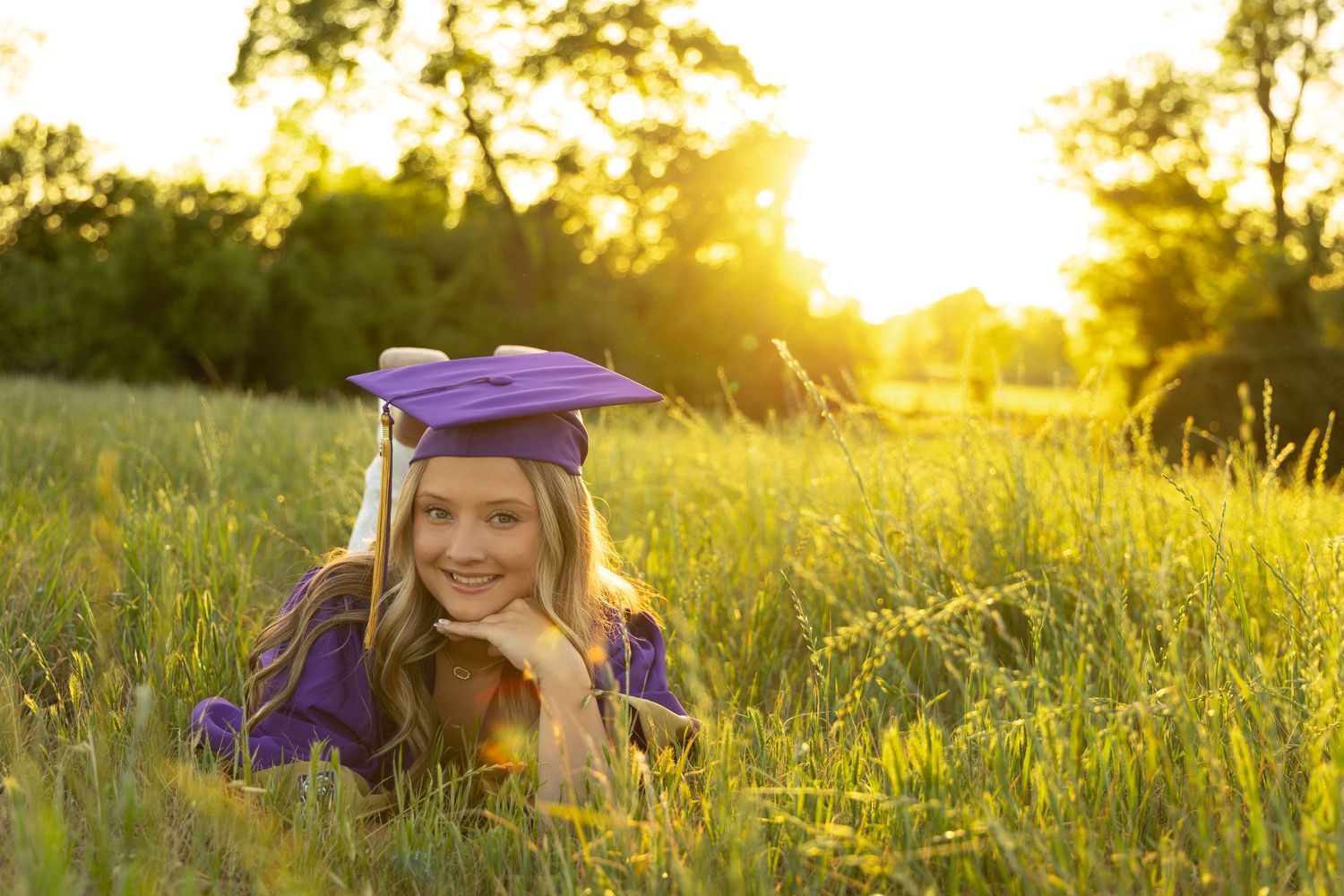 Smiling graduate in purple cap and gown lying in green field at golden sunset.