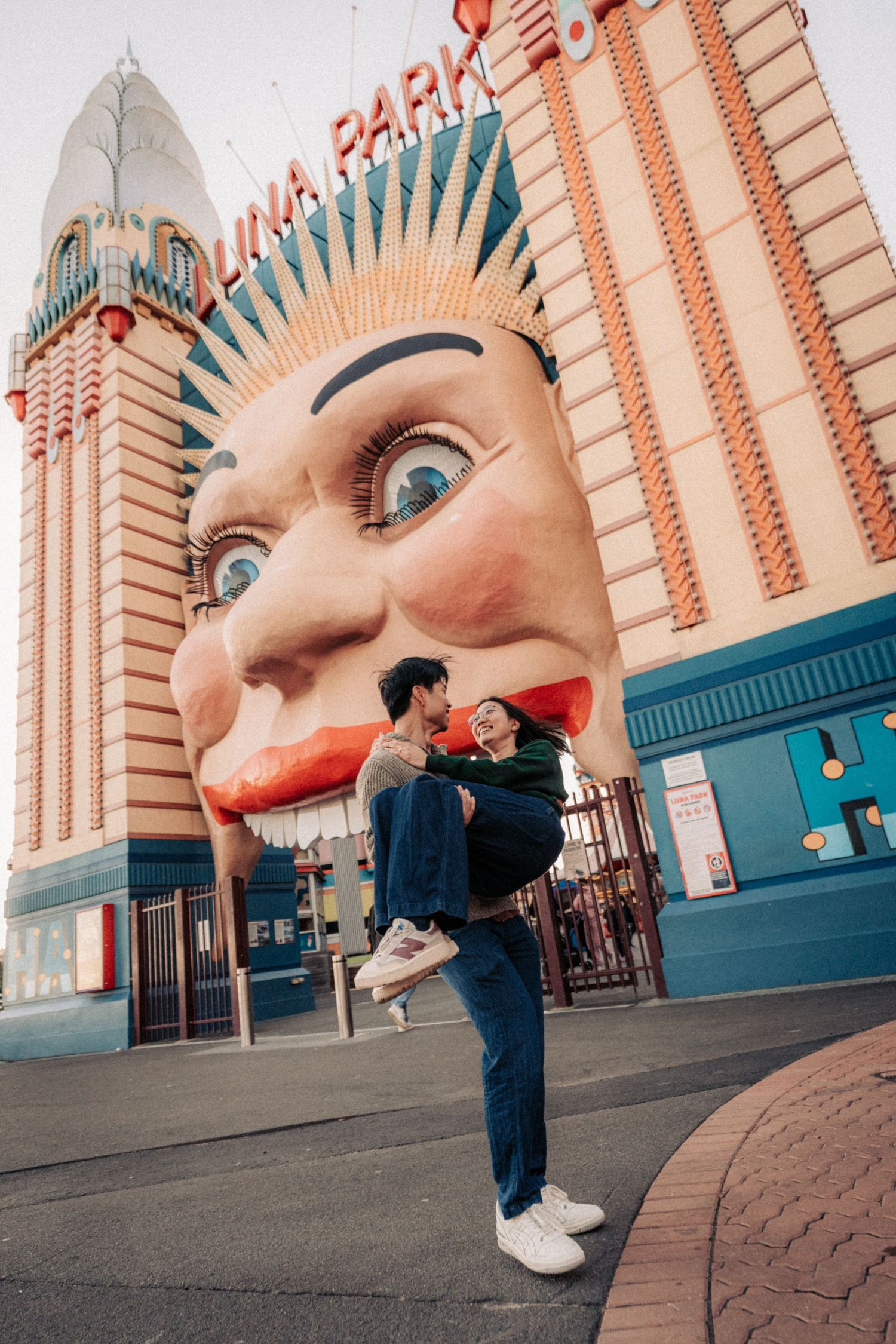 Couple embracing at Luna Park Sydney entrance, featuring the iconic giant face facade, in a romantic outdoor portrait.