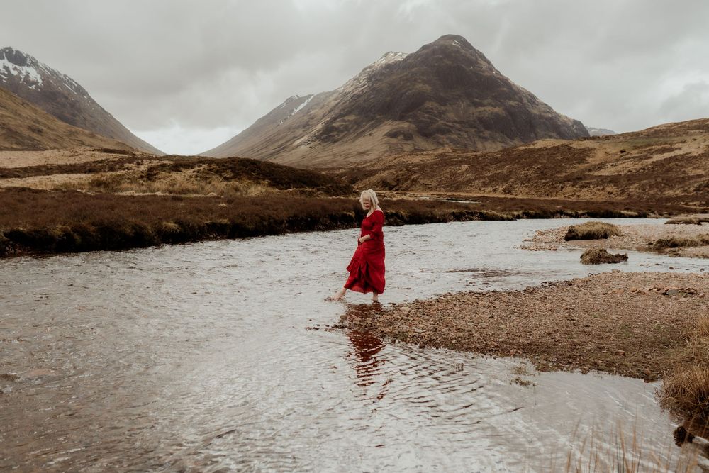 Woman in red dress standing in a river with dramatic Scottish Highland mountains in the background.