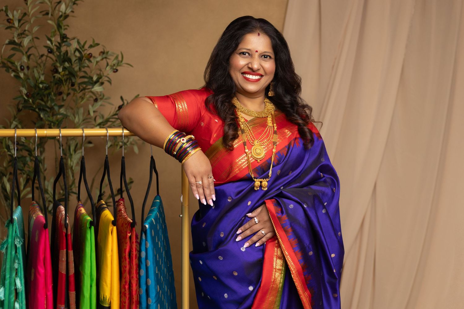 Woman in vibrant blue and red silk saree with gold jewelry poses confidently beside colorful garments on a rack.