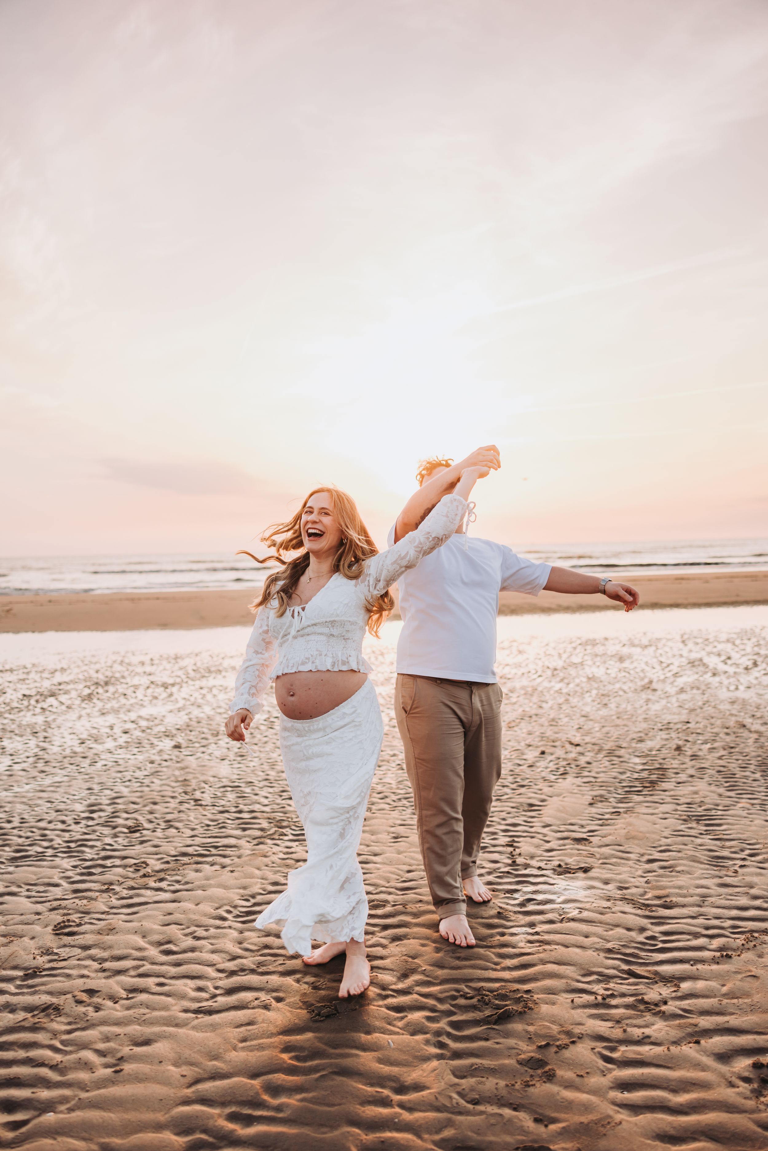 Golden light warm edited image of joyful pregnant couple dancing barefoot on a sandy beach at sunset, woman in white lace dress laughing.