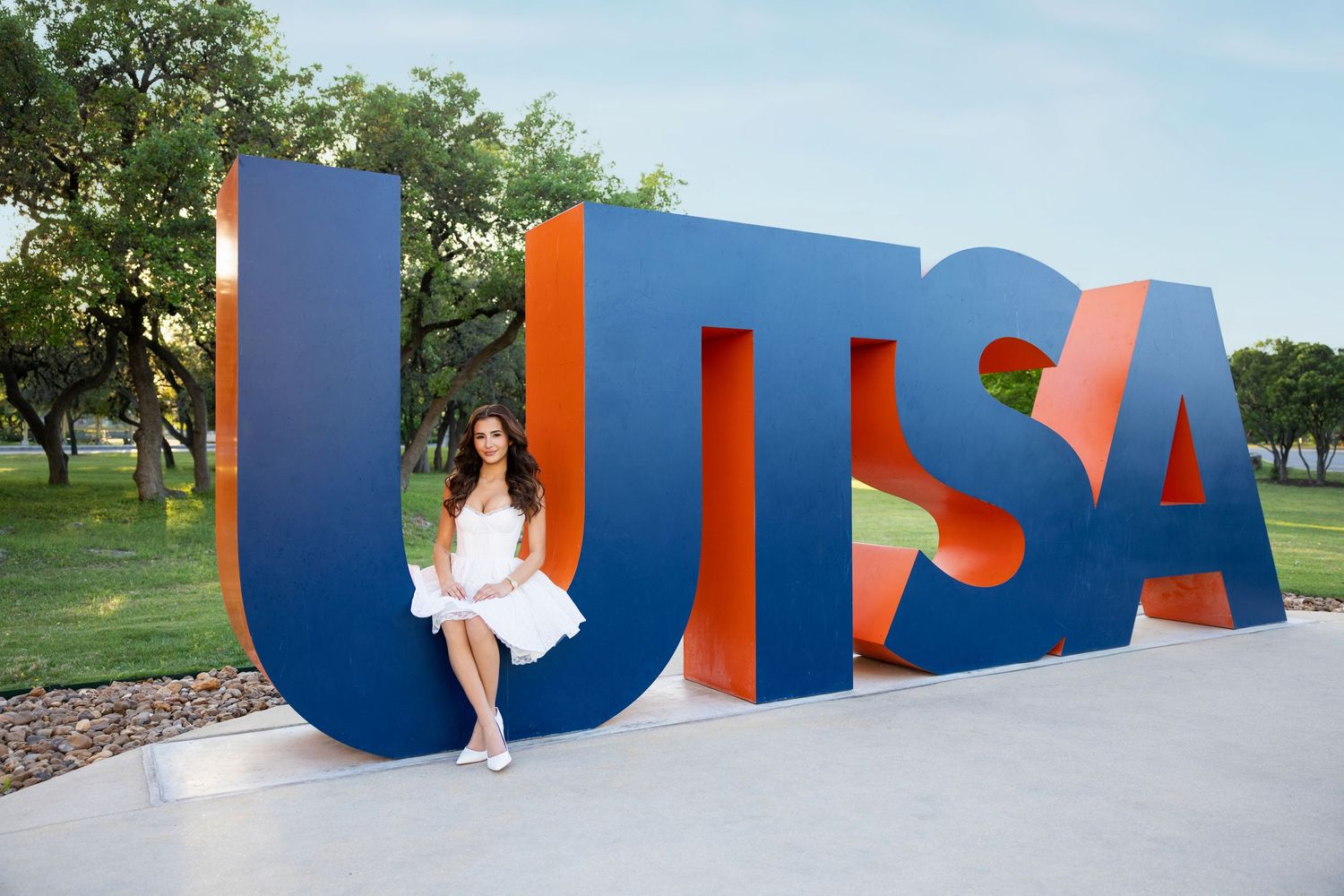 College graduate poses in white dress holding cap before large UTSA letters outdoors on sunny day.