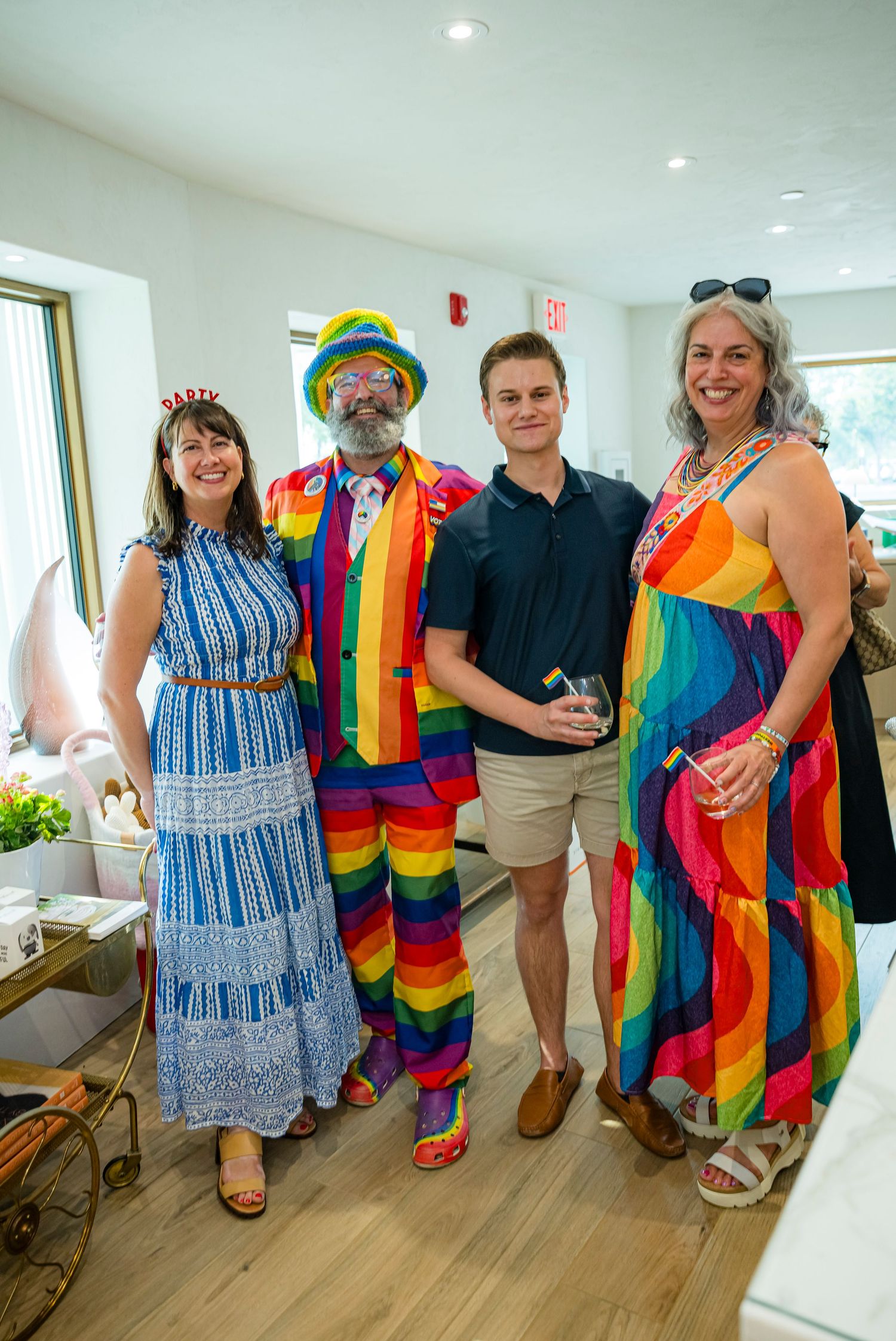 Four people posing indoors, two wearing vibrant rainbow outfits, at a colorful pride-themed celebration event.