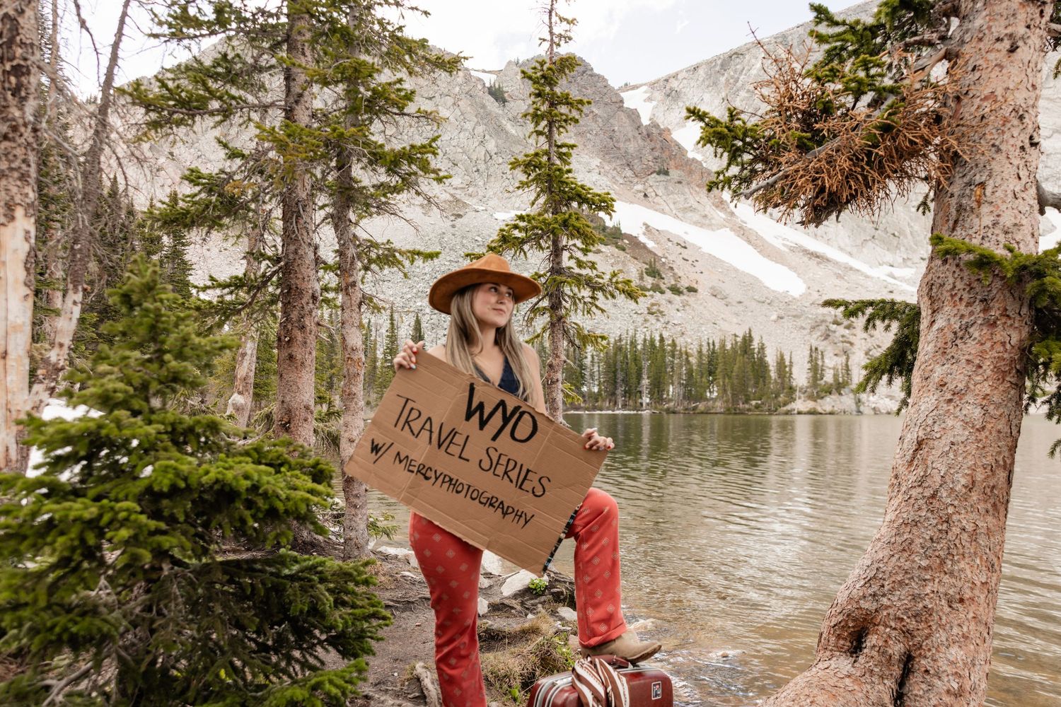 Hiker holding a wooden sign beside a mountain lake surrounded by pine trees and rocky peaks.