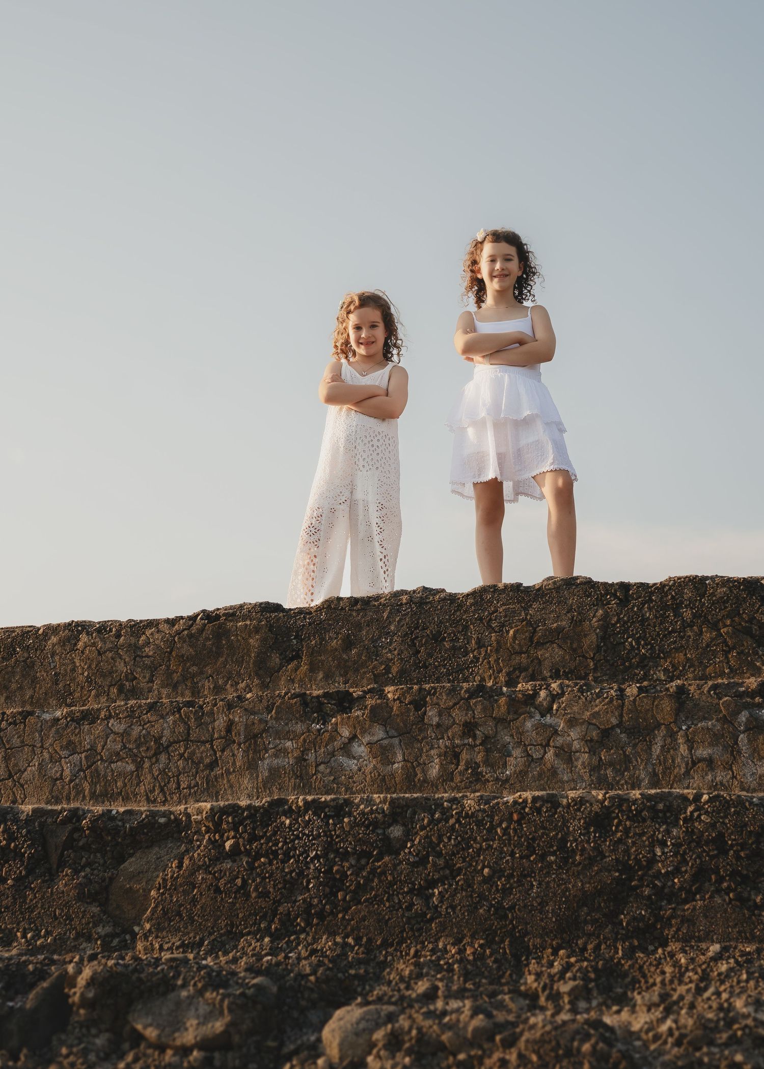Two women in dresses pose on rocky coastal ruins overlooking a calm bay at golden hour.