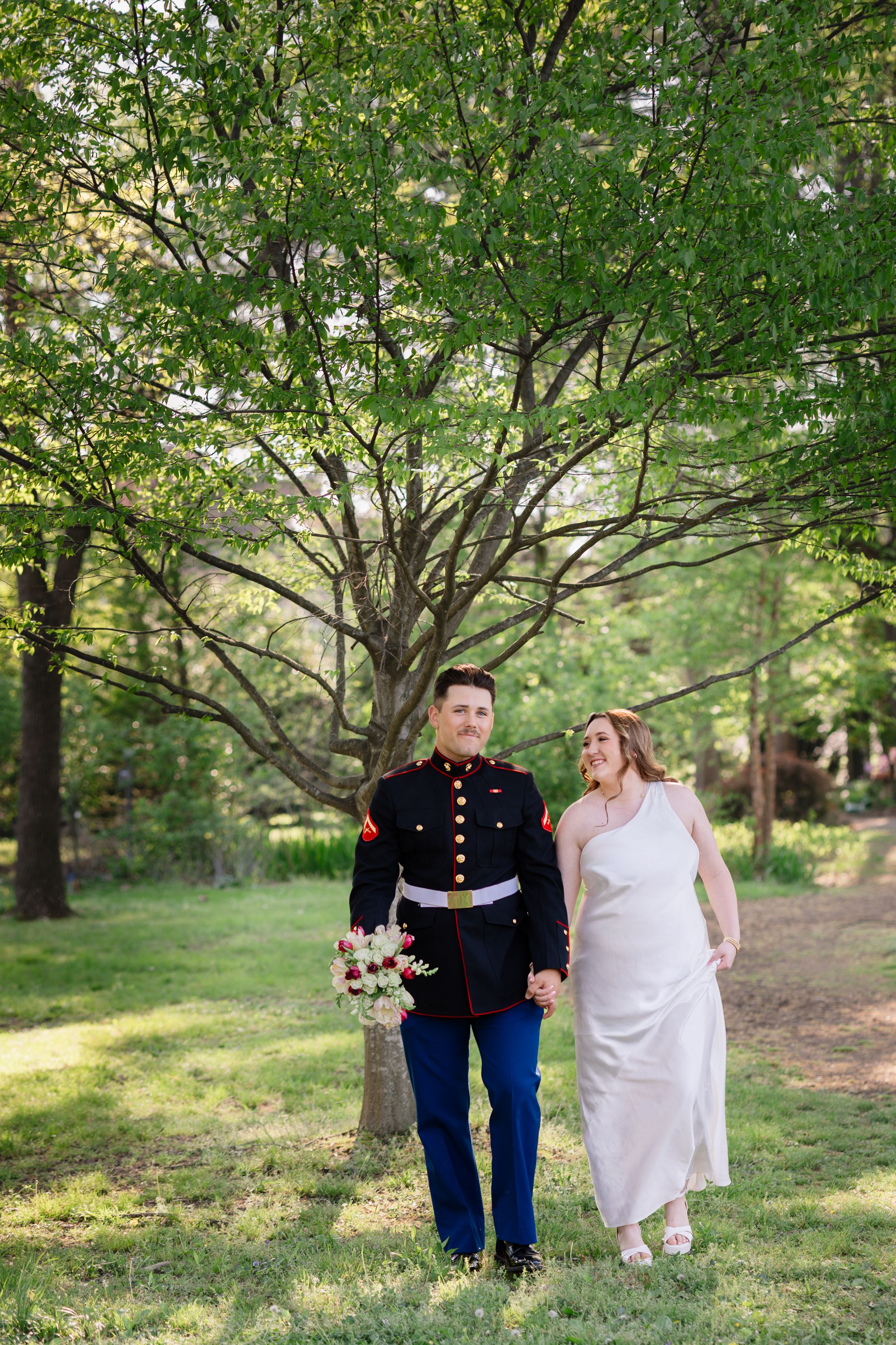 A Marine in dress uniform walks hand-in-hand with a bride in a white gown through a lush green park on their wedding day.