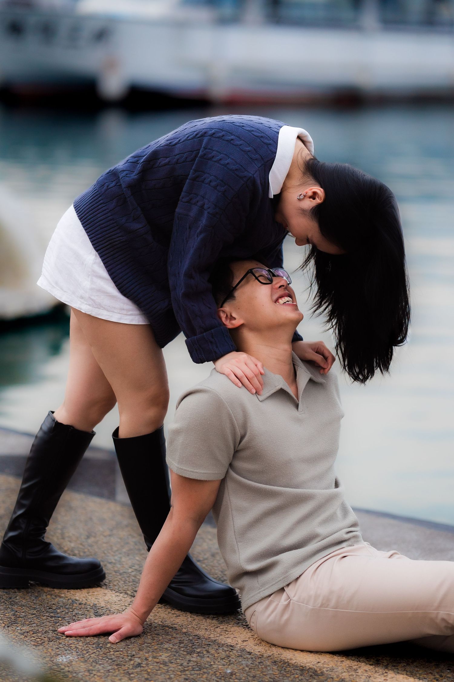 Couple sharing a joyful moment by the waterfront, woman in navy sweater leaning over smiling man seated on ground.