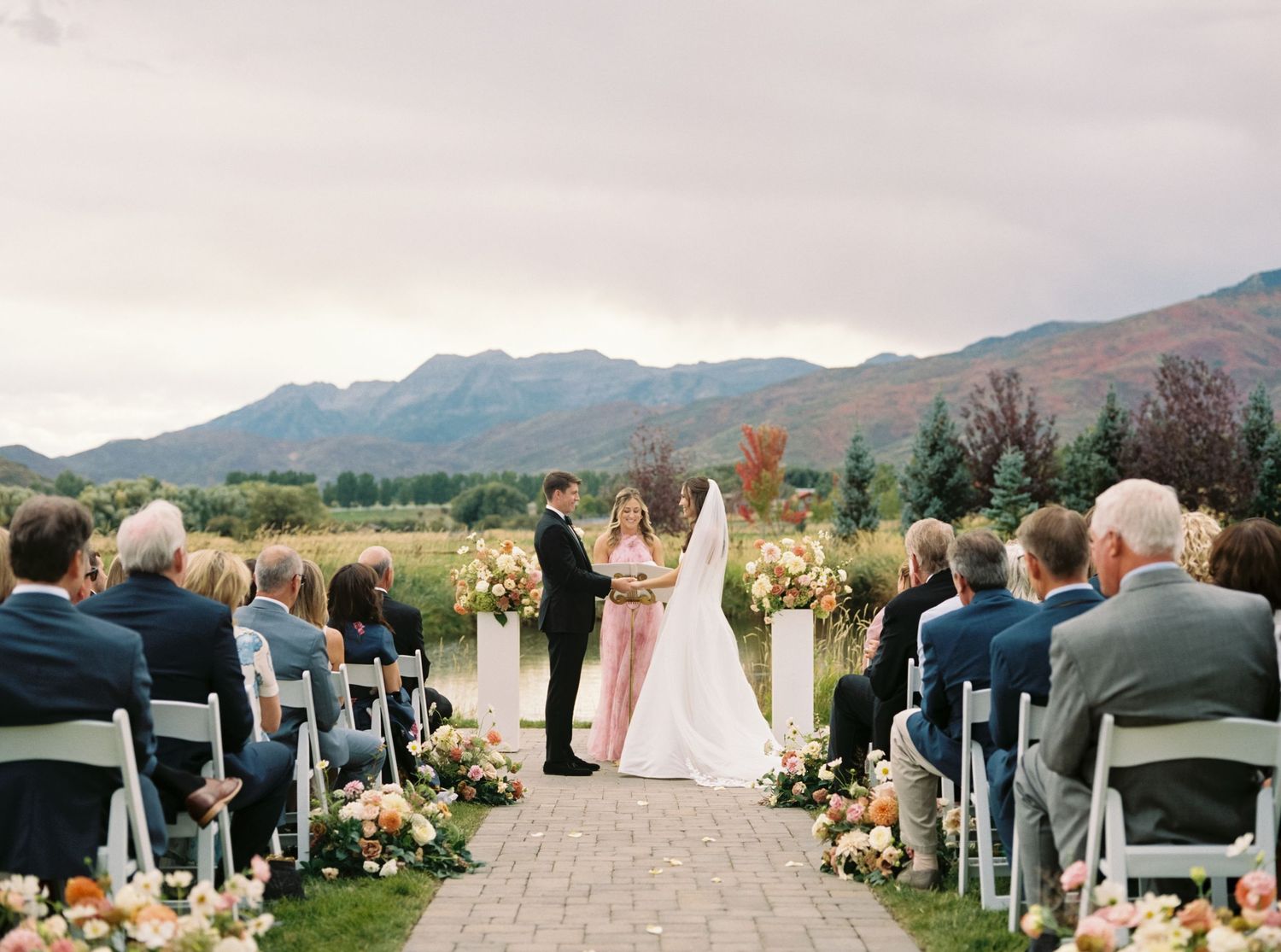 Outdoor wedding ceremony with mountain backdrop, floral arch, bride and groom at altar, guests seated on white chairs.