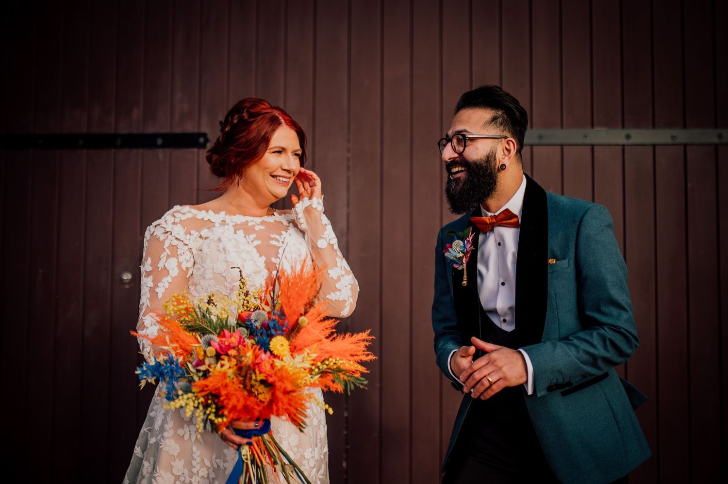 A couple in wedding attire shares intimate moments in front of a dark wooden wall with vibrant orange florals. Worcestershire Wedding photog