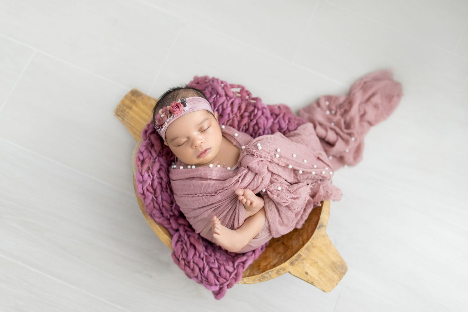 Newborn baby sleeping peacefully in wooden bowl with pink knit blanket and floral headband.