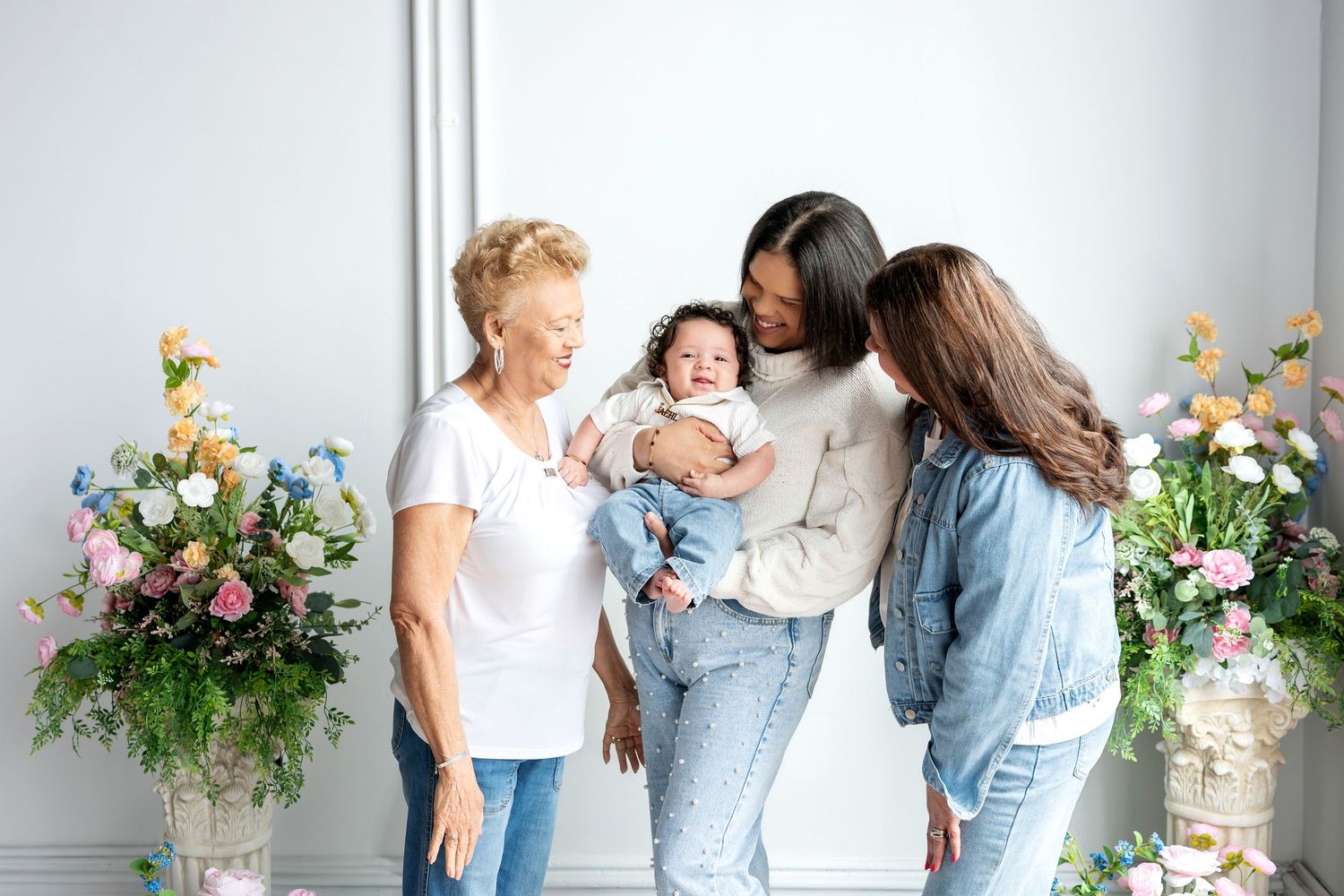 Three generations of women admire a smiling baby in a bright floral studio setting.