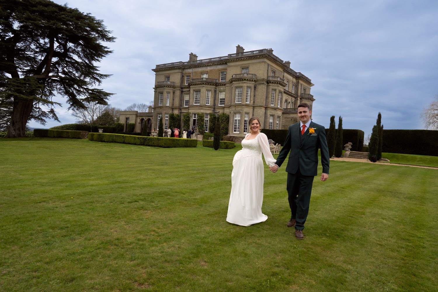 Bride and groom holding hands on lush green lawn in front of grand historic manor house wedding venue.