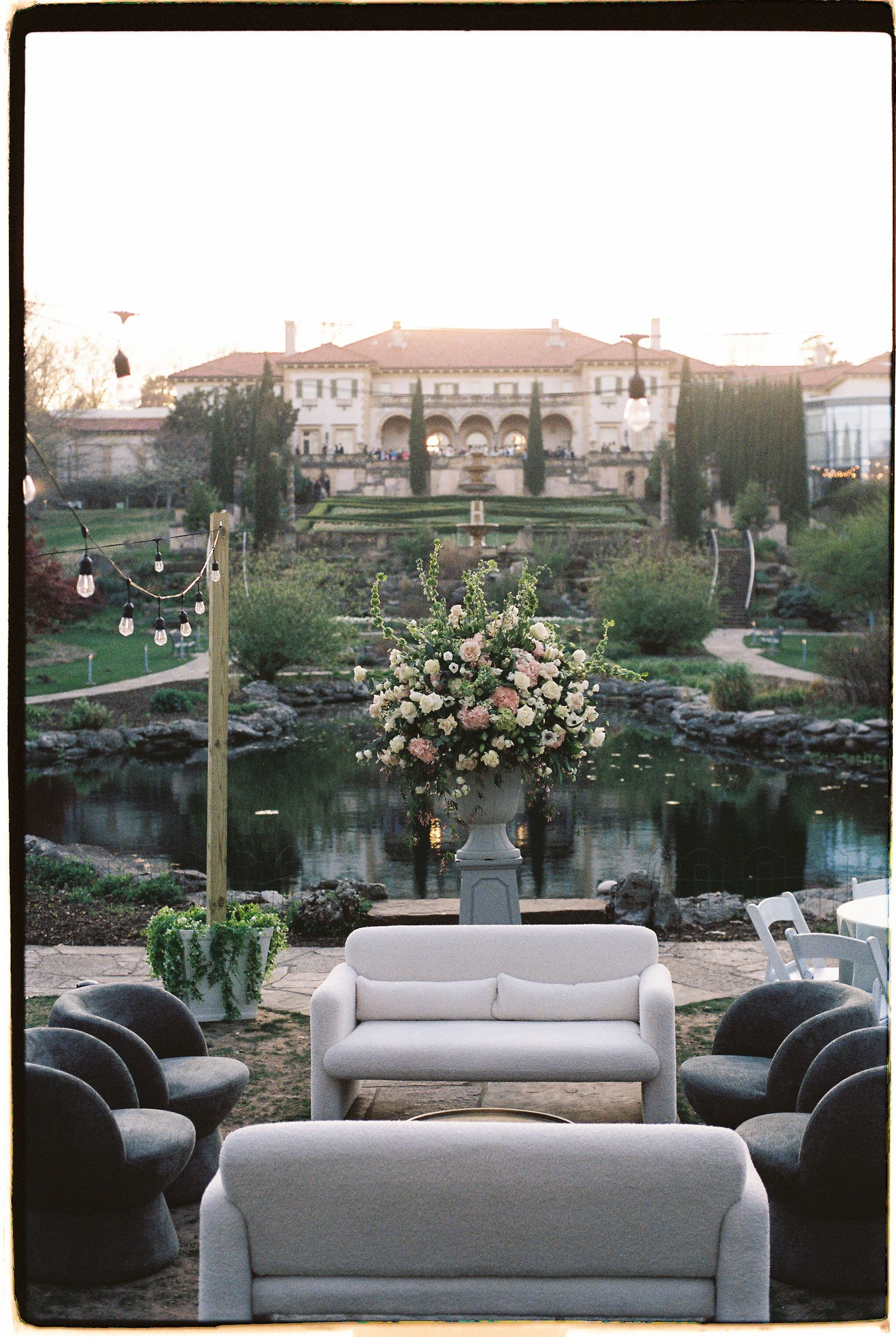 Elegant outdoor wedding lounge with gray sofas, floral arrangement, pond, and grand estate backdrop at sunset.