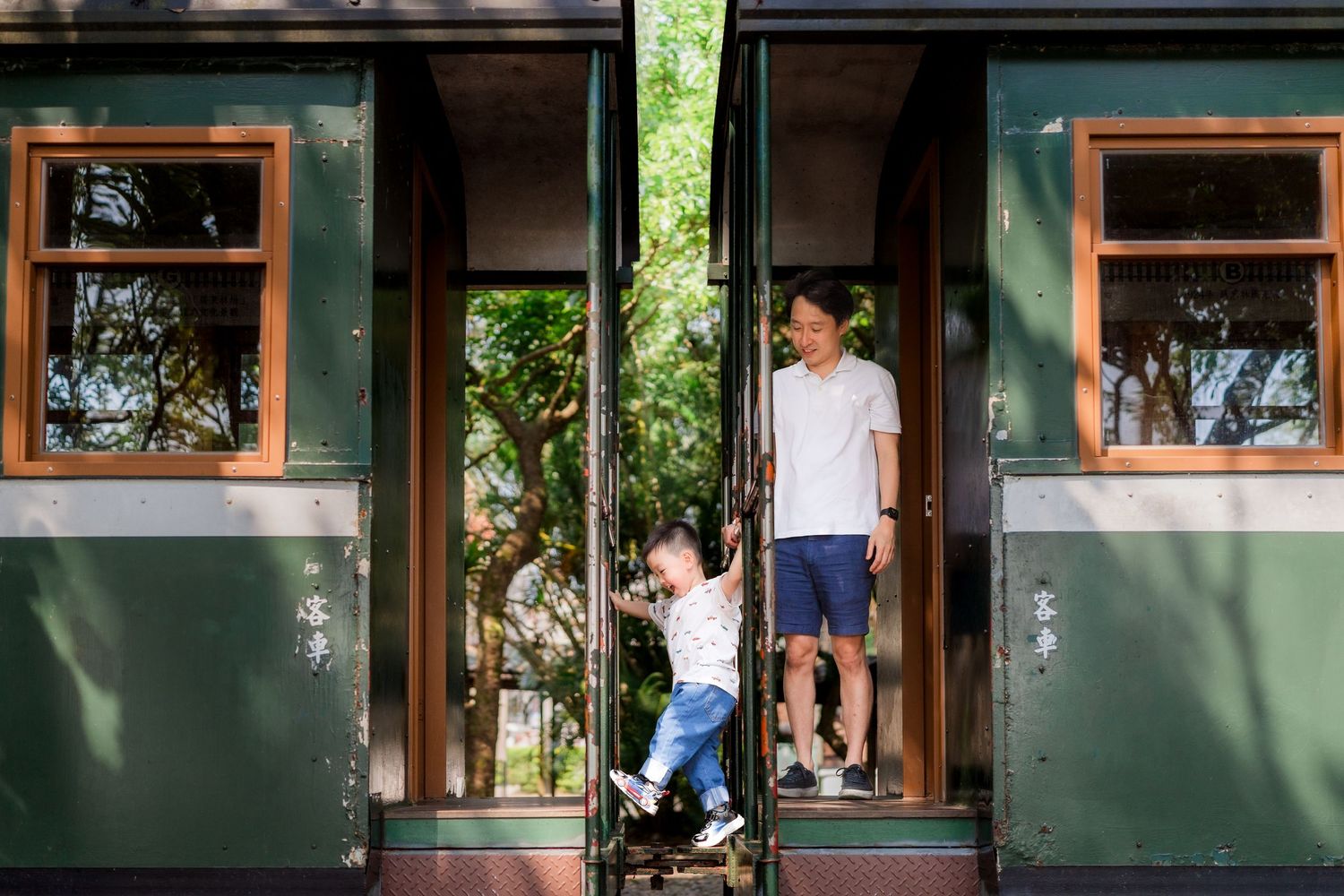 Father and toddler exploring a vintage green train car doorway surrounded by lush trees.