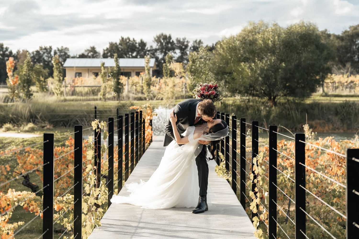 Bride and groom share a romantic dip kiss on a bridge surrounded by autumn foliage near a serene lake.