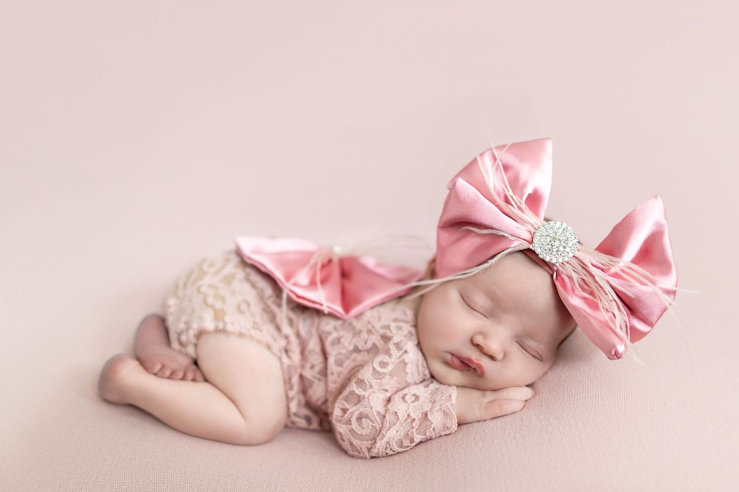 Sleeping newborn baby girl in pink lace romper and large satin bow headband on soft pink background.