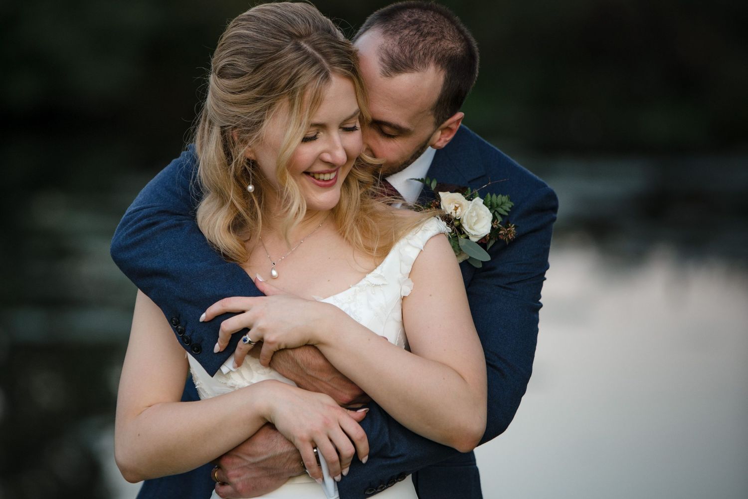 A groom in a navy suit embraces his smiling bride in a white dress near a serene lakeside backdrop.