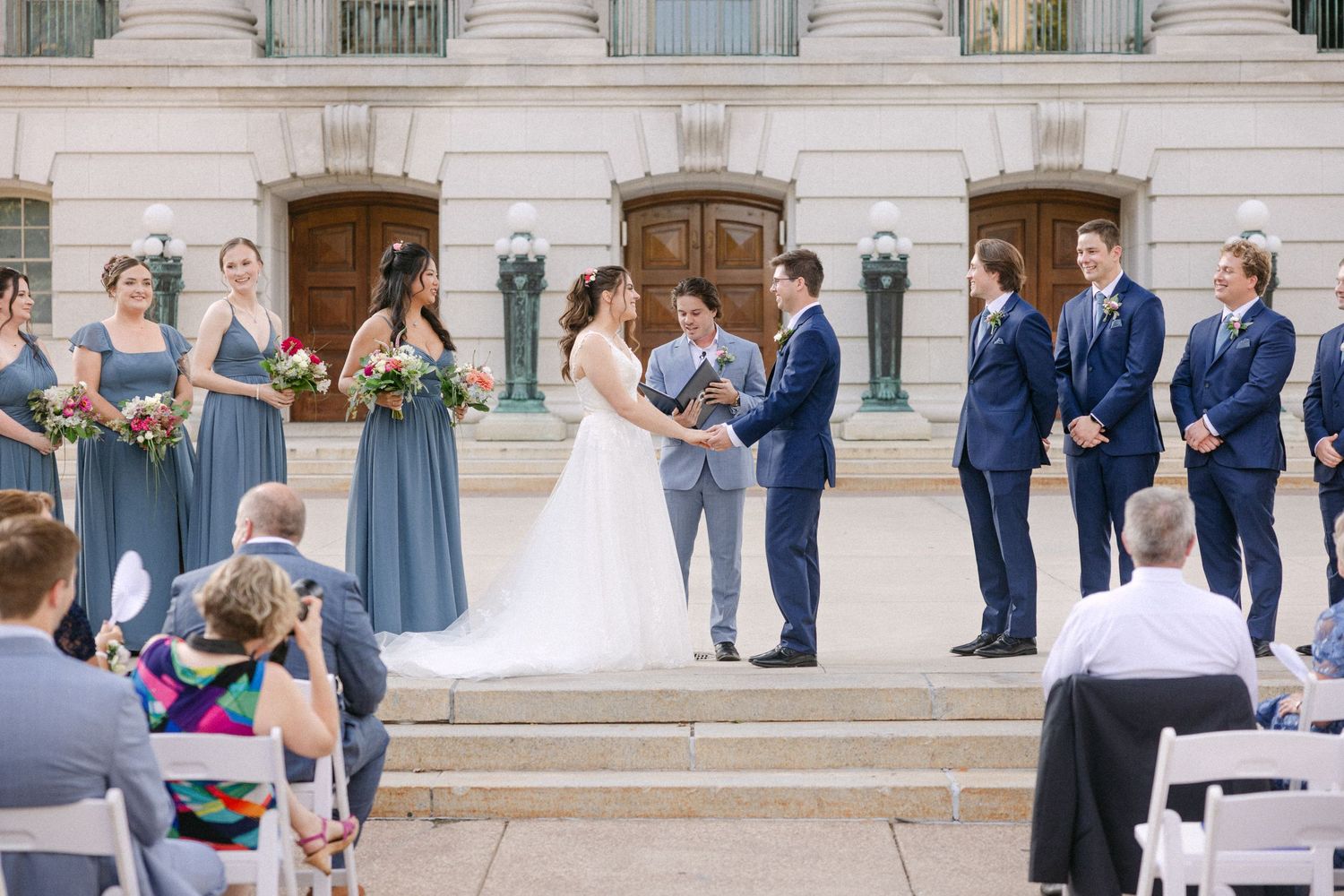 Outdoor wedding ceremony on stone steps with bride and groom exchanging vows, bridesmaids in blue dresses, groomsmen in navy suits.