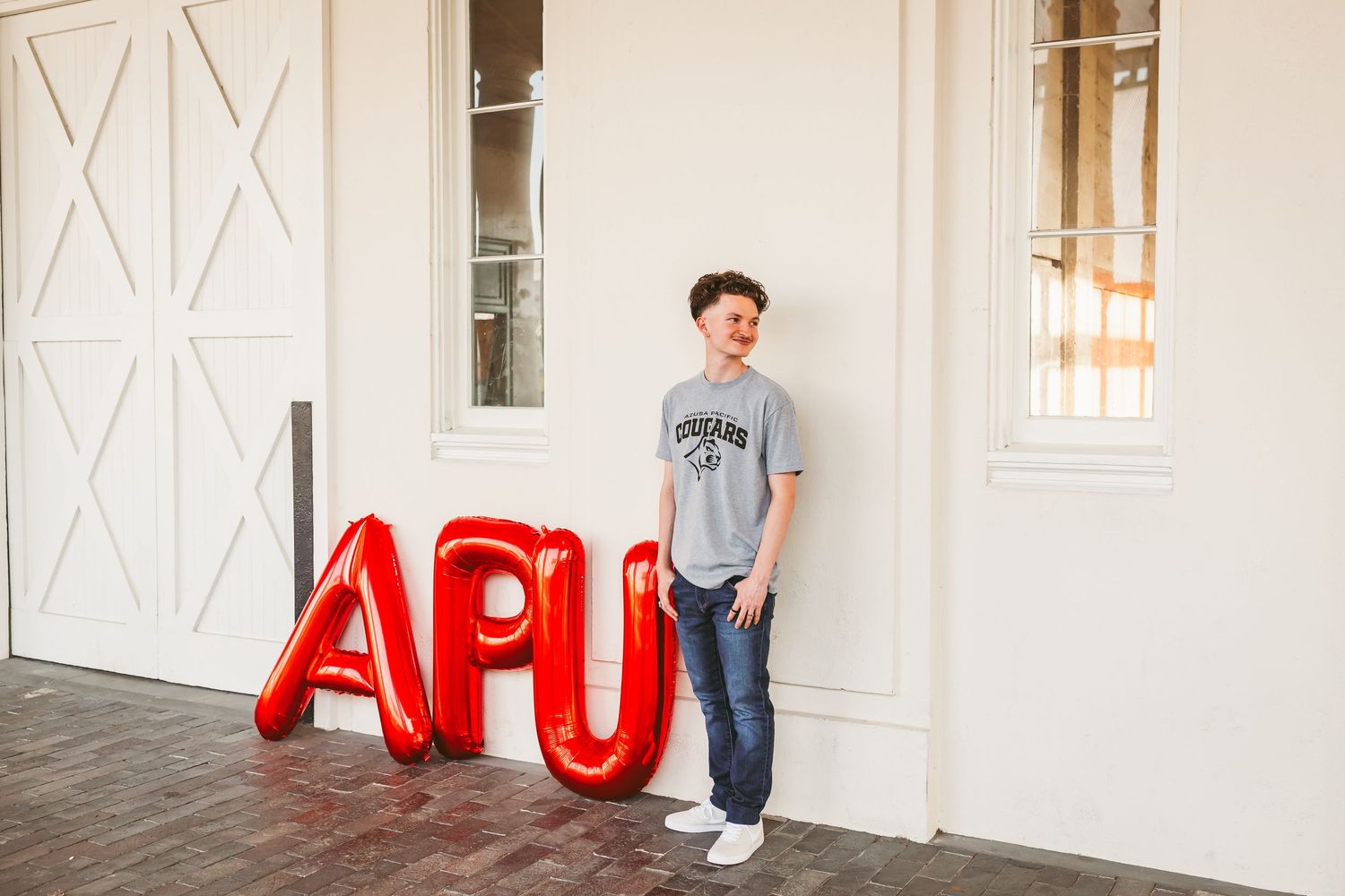 Young man posing beside red APU letter balloons outside a white building, wearing a gray college t-shirt and jeans.
