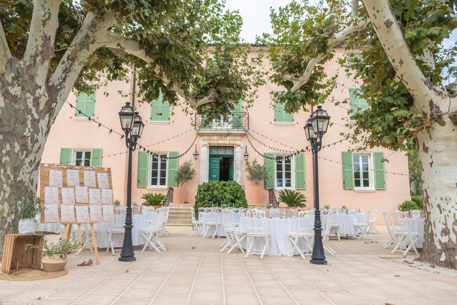 A serene outdoor courtyard with pink stucco walls, potted plants, and white chairs arranged for an elegant event setup.
