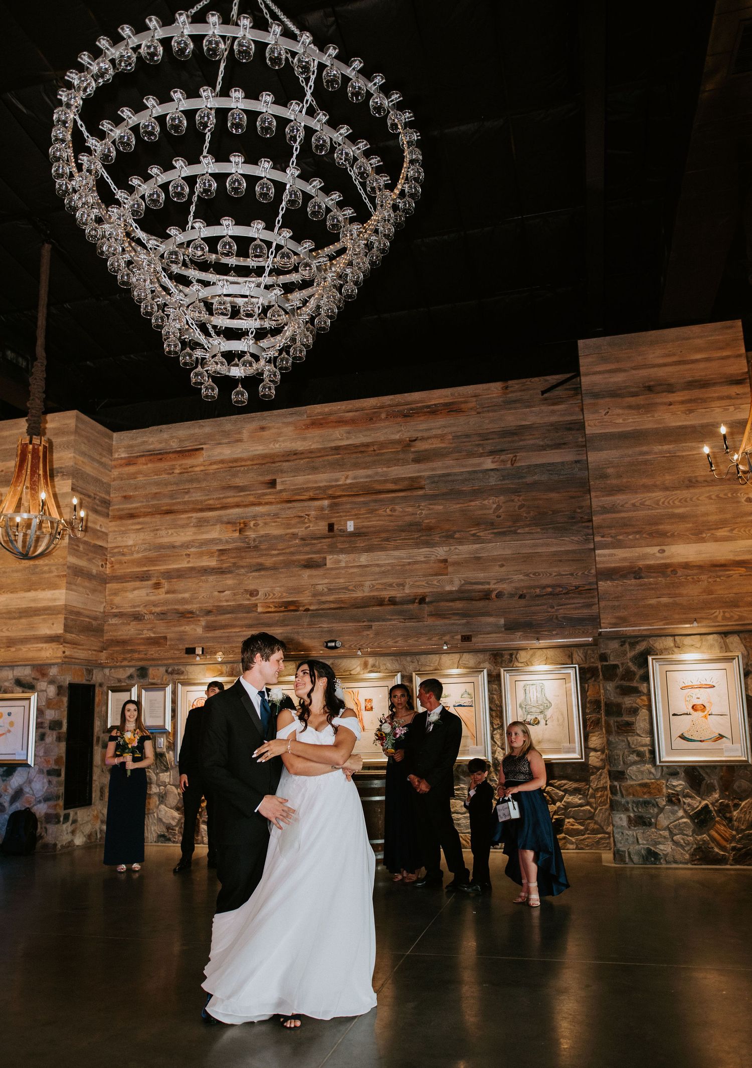 A couple dances in a rustic barn venue with elegant crystal chandeliers hanging overhead during their wedding reception.