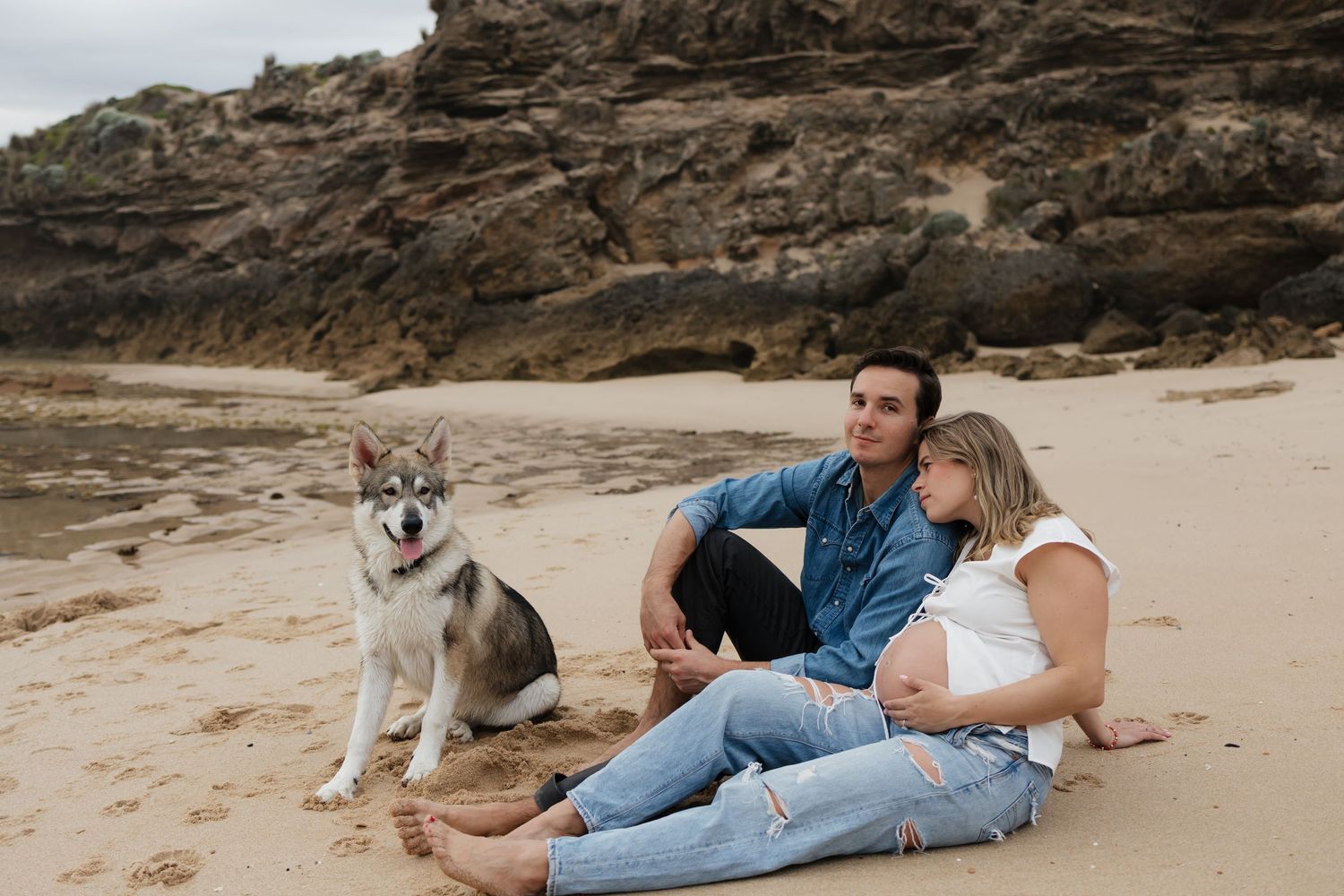 Pregnant couple sitting on sandy beach with Husky dog, rocky cliffs in background, casual maternity photo shoot.