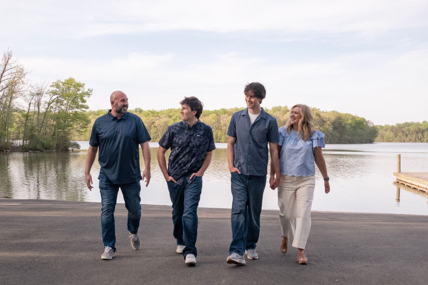 Four people walking together near a lakeside dock on a sunny day, surrounded by lush green trees.
