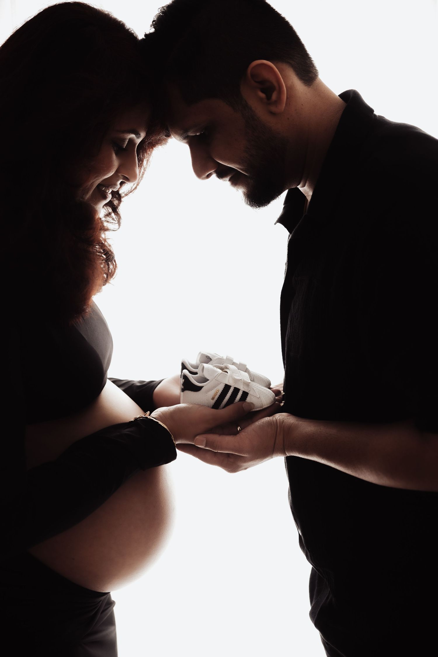 Silhouette of pregnant couple holding tiny baby shoes together, sharing an intimate moment in soft backlit photography.