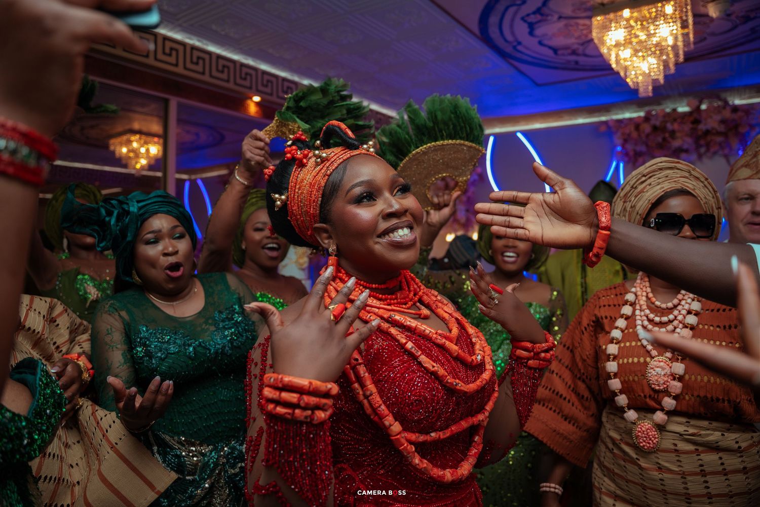 Joyful bride in traditional Nigerian attire with coral beads and green feathered headpiece celebrates at vibrant wedding ceremony.