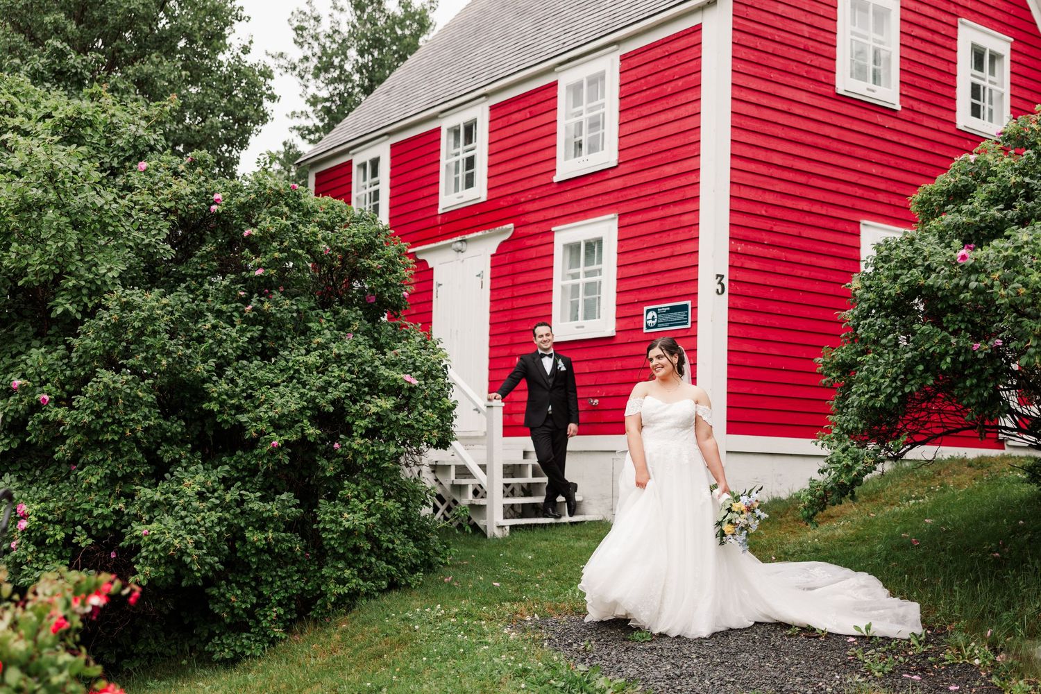 Bride and groom pose outside a vibrant red historic building surrounded by lush greenery on their wedding day.