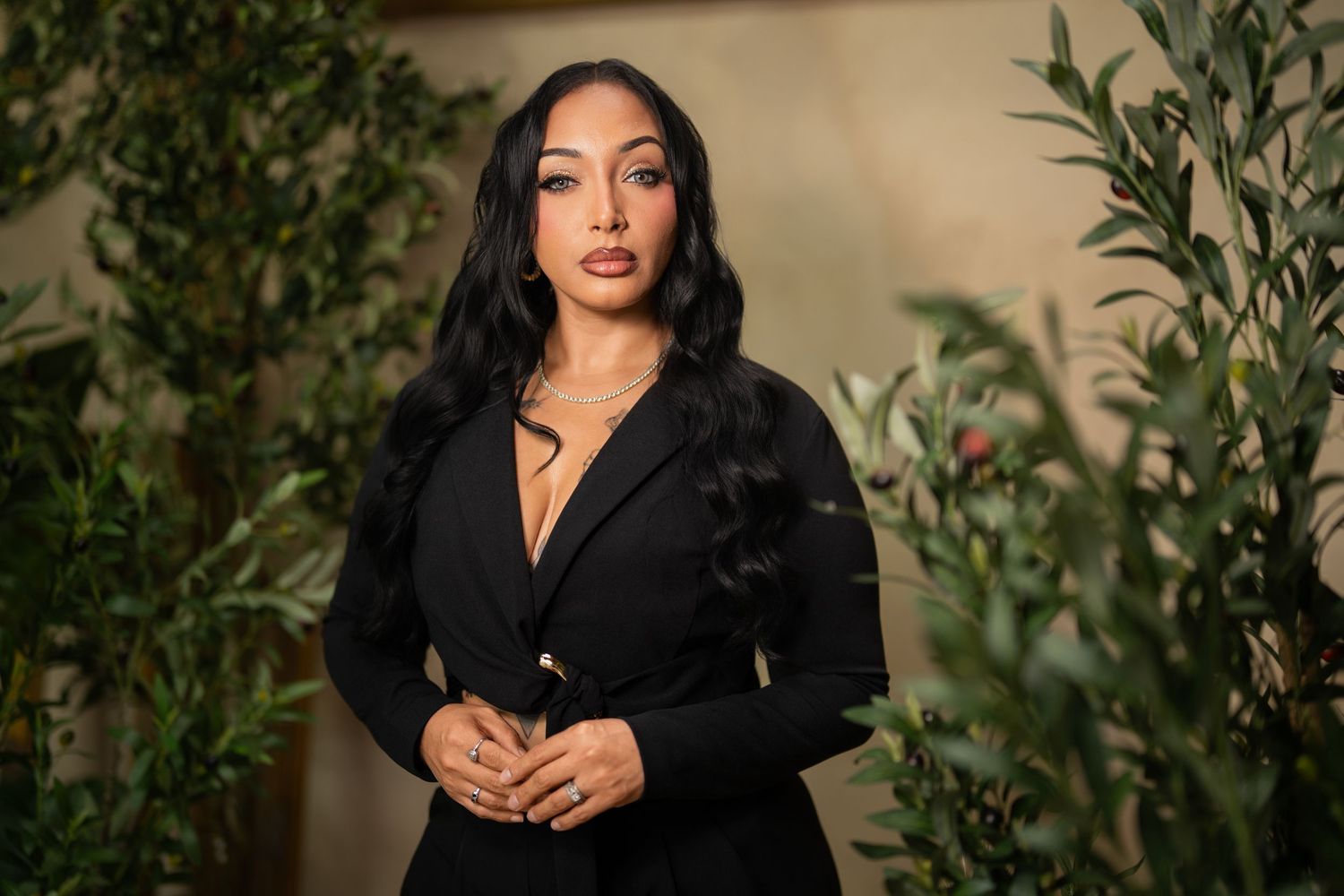 Woman in black blazer dress poses among green plants, featuring long dark hair and elegant style.