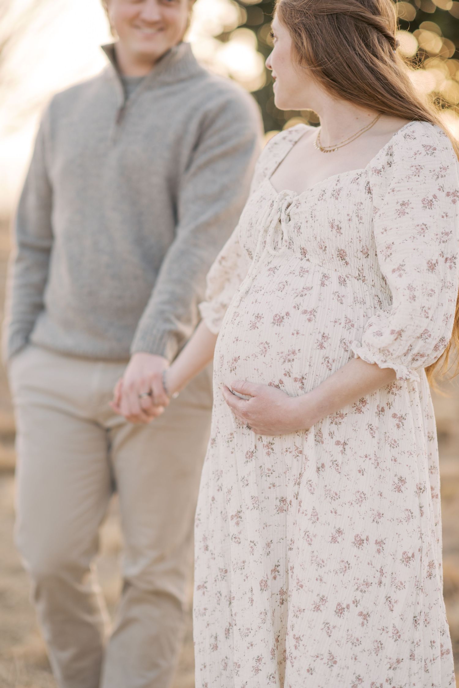 Pregnant woman in floral dress holds baby bump while holding hands with man in gray sweater outdoors.