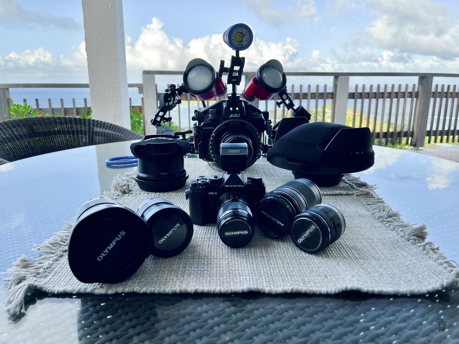 Sunglasses and camera equipment arranged on a stone ledge against a bright blue ocean and sky backdrop.