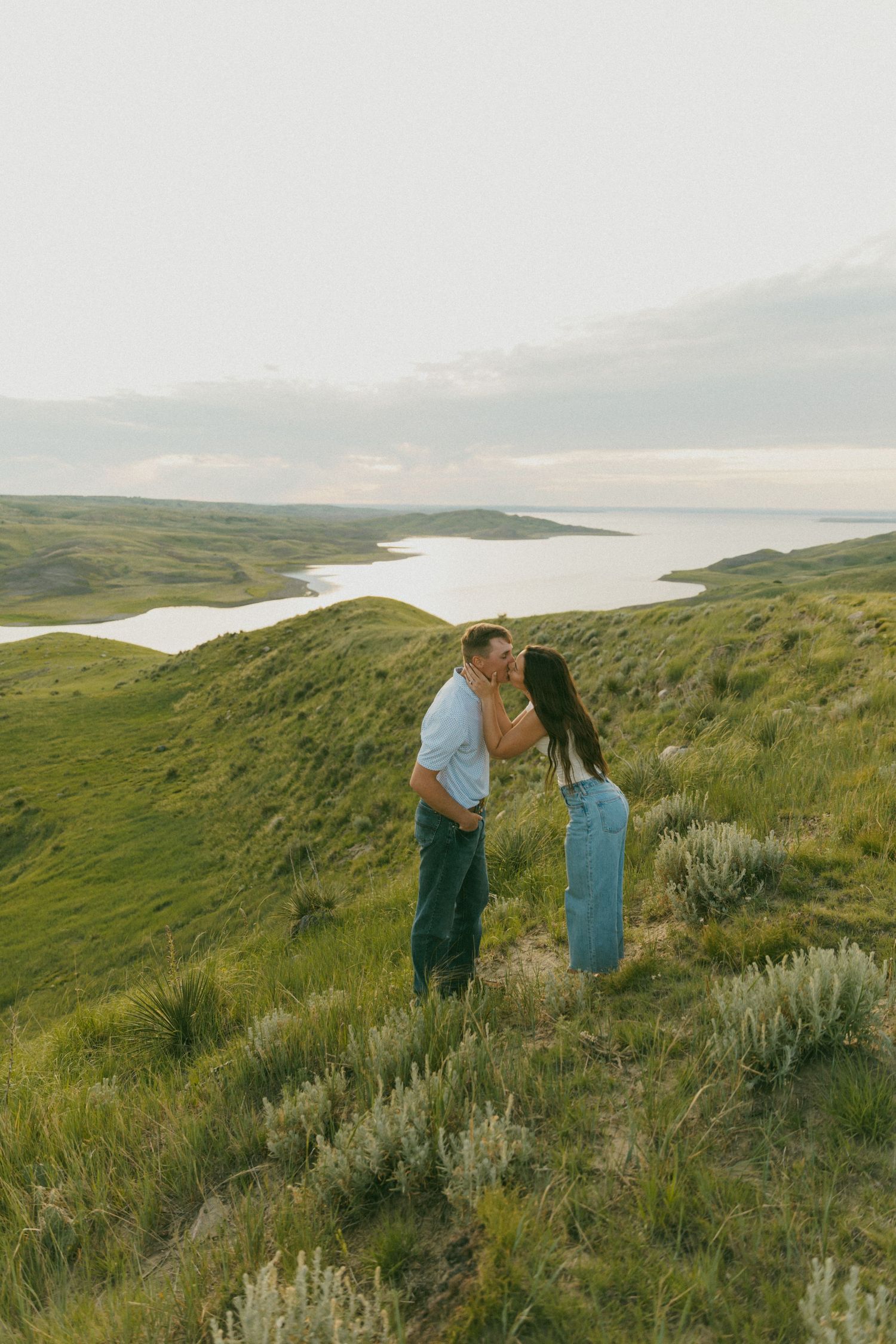 A couple shares a romantic moment on a hilltop overlooking a scenic lake and rolling hills.