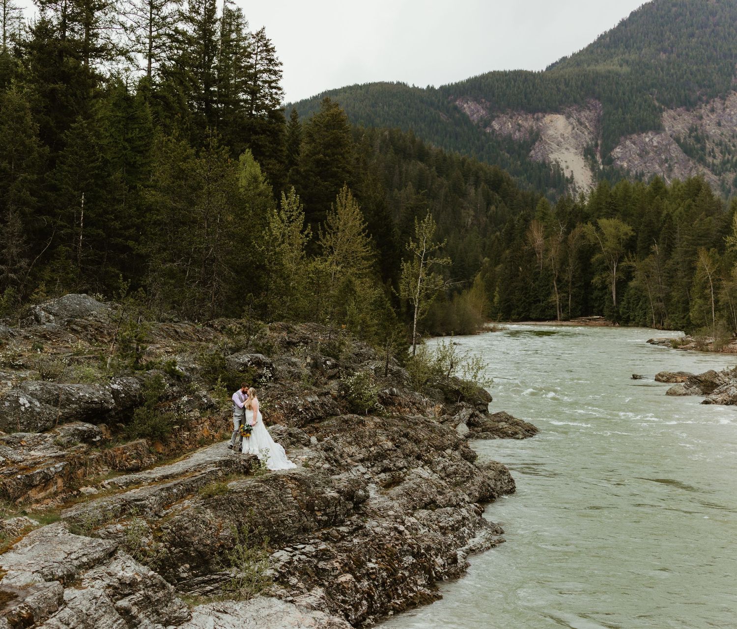 Bride and groom stand on rocky riverbank with turquoise water, surrounded by lush forest and mountains.