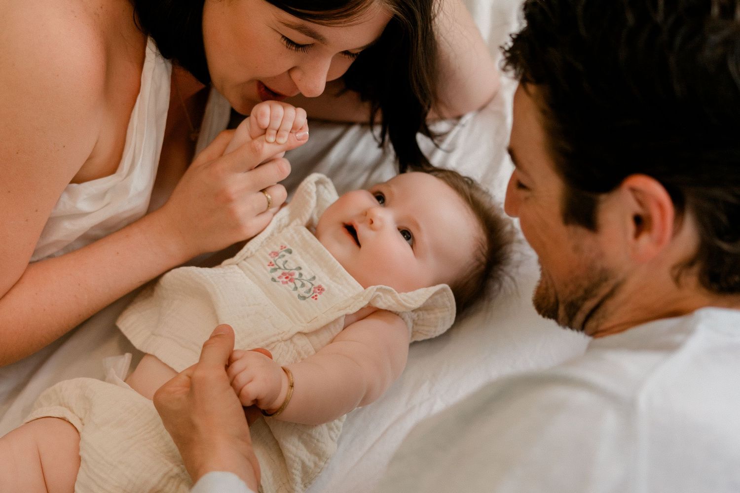Parents lovingly interact with their baby lying on a white bed in a warm family lifestyle photography session.
