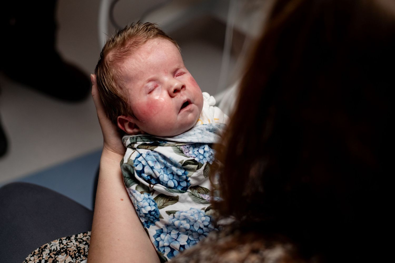 A baby wearing a blue and white patterned outfit is being held against a dark background.