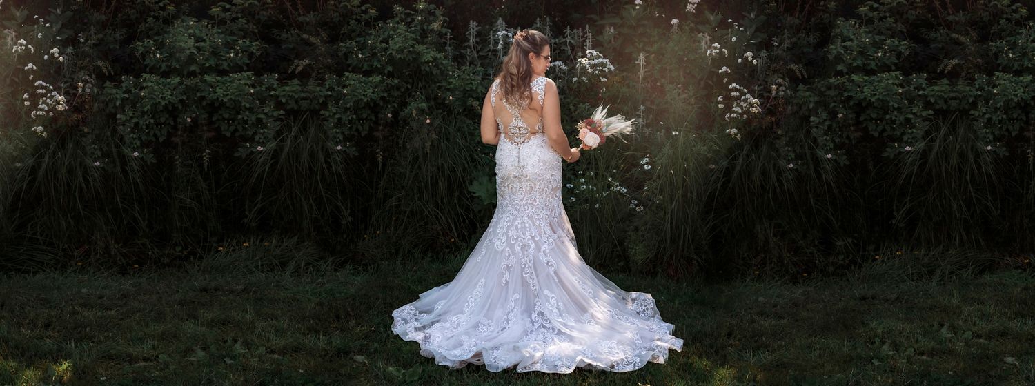 Bride in lace mermaid wedding gown holding bouquet stands before lush dark forest with sunlight streaming through.