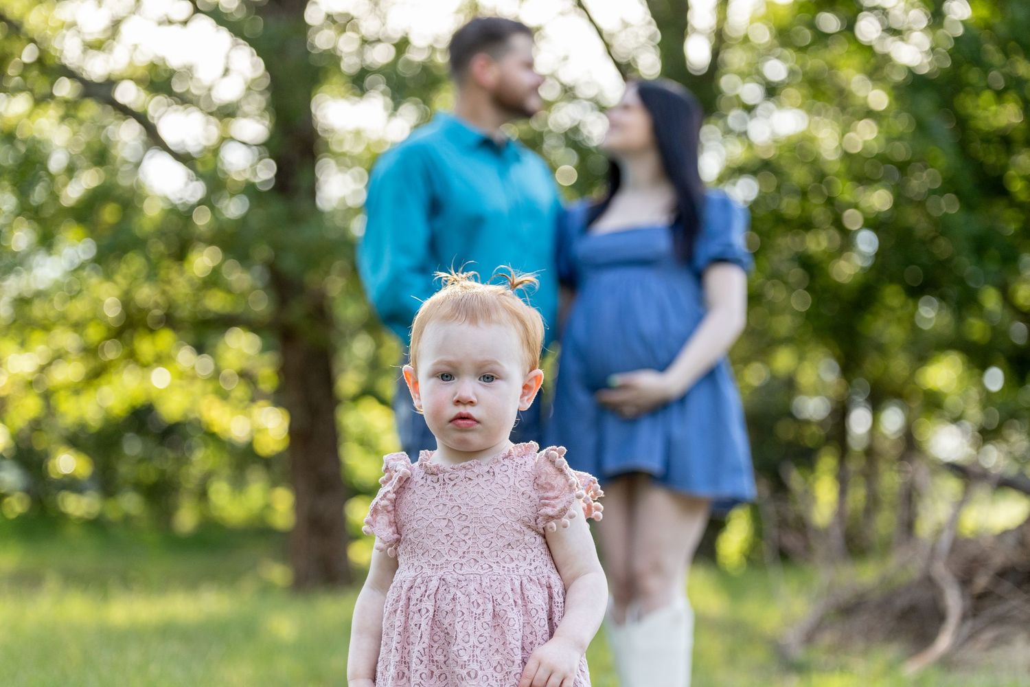 A toddler girl in a pink dress stands in focus outdoors while her pregnant mother and father share a moment behind her.