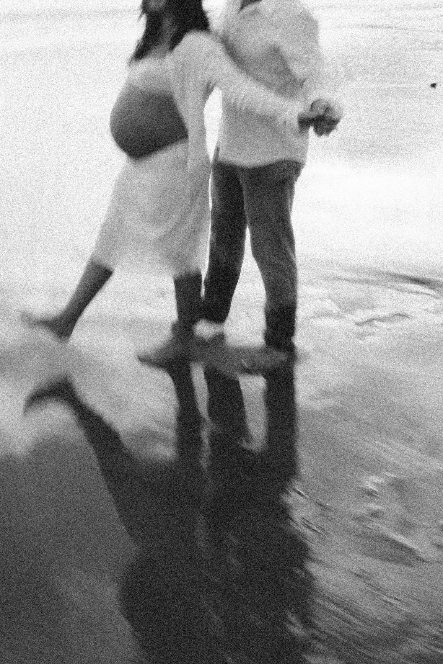 Black and white photo of a pregnant couple embracing on a wet beach, their reflections visible in the water.