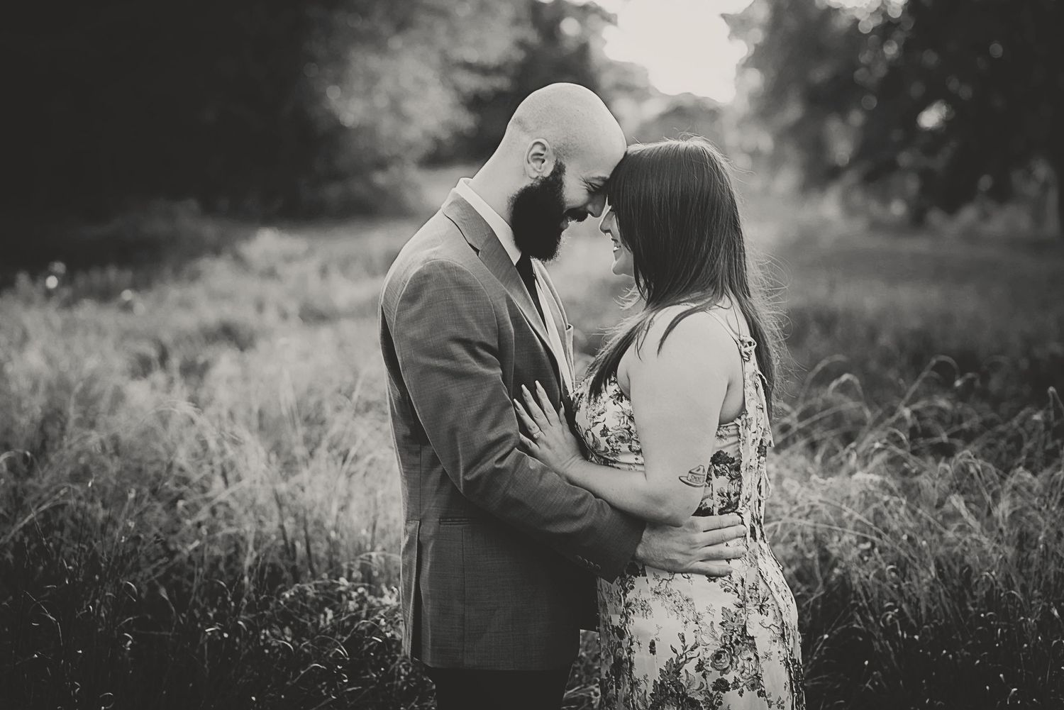 Black and white photo of a couple embracing in a field, sharing a romantic moment surrounded by tall grass and trees.