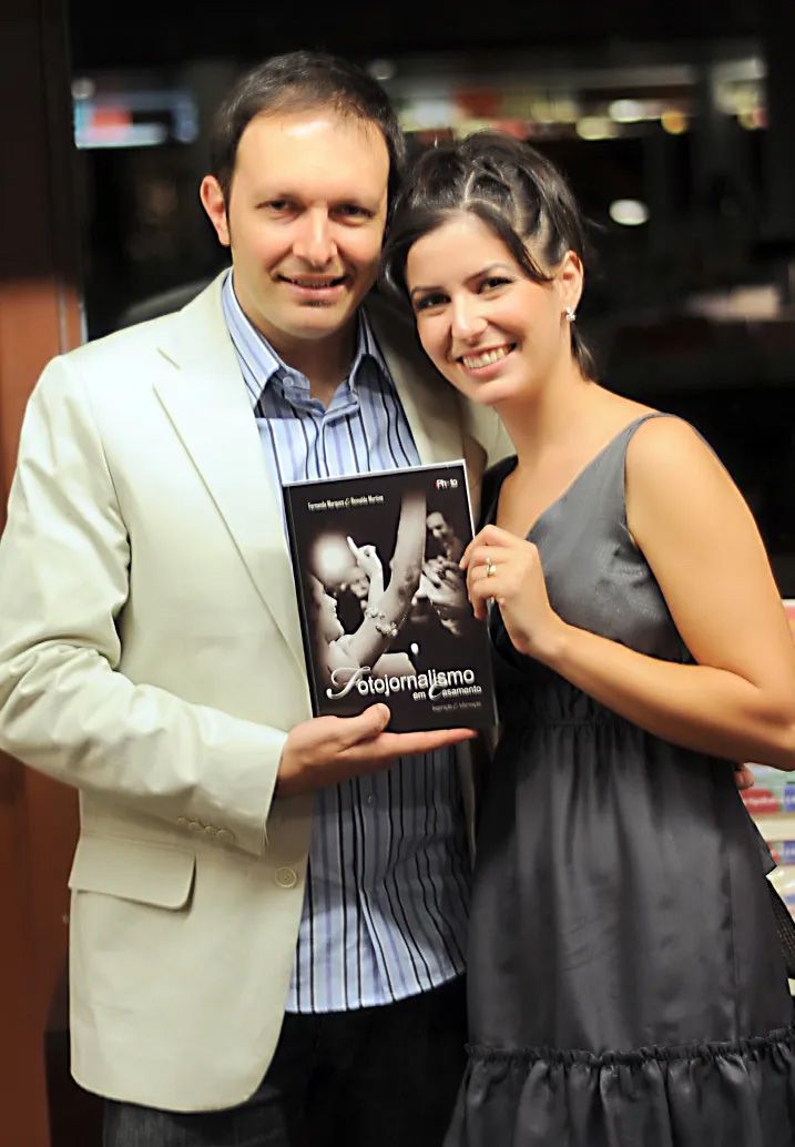 A man and woman smiling while holding a book titled Fotojornalismo at what appears to be a book launch event.