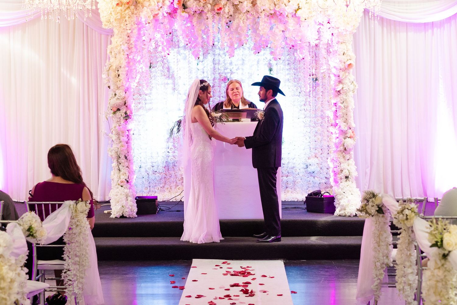 Bride and groom hold hands at wedding altar with floral backdrop, officiant behind them, groom wearing cowboy hat.